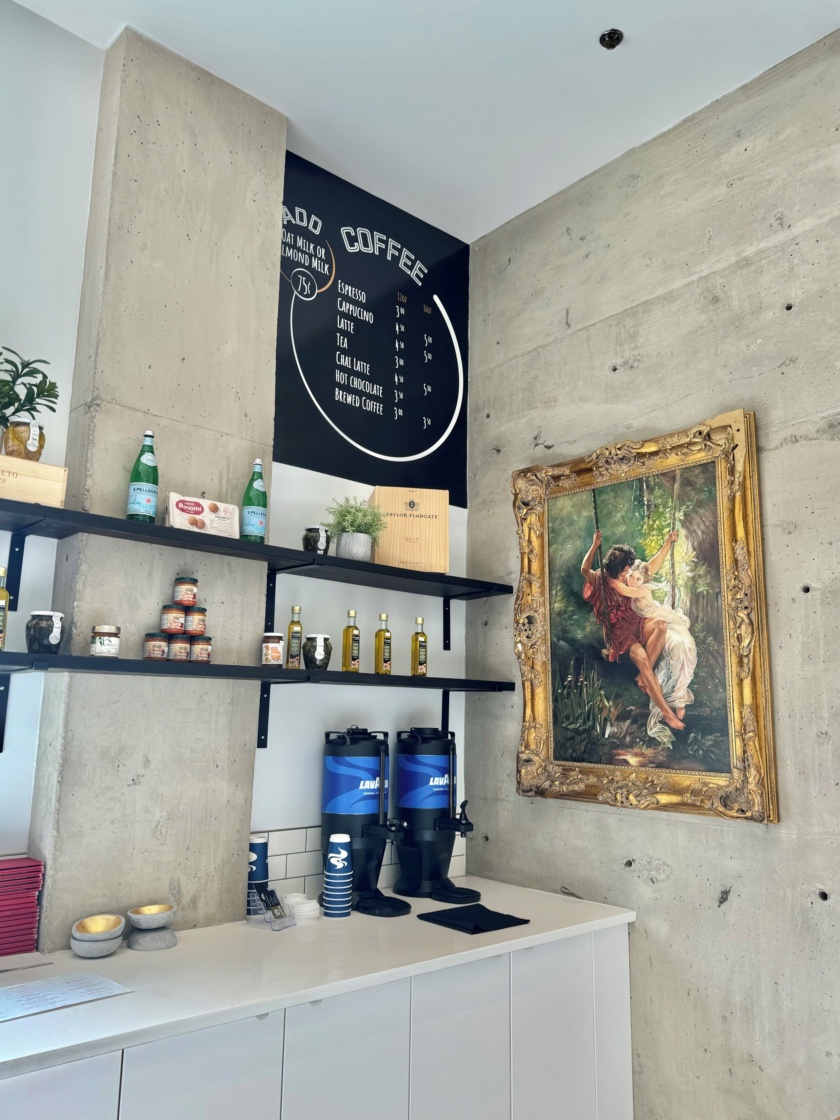 Interior de café com quadro de cardápio, prateleiras com temperos e uma pintura emoldurada.