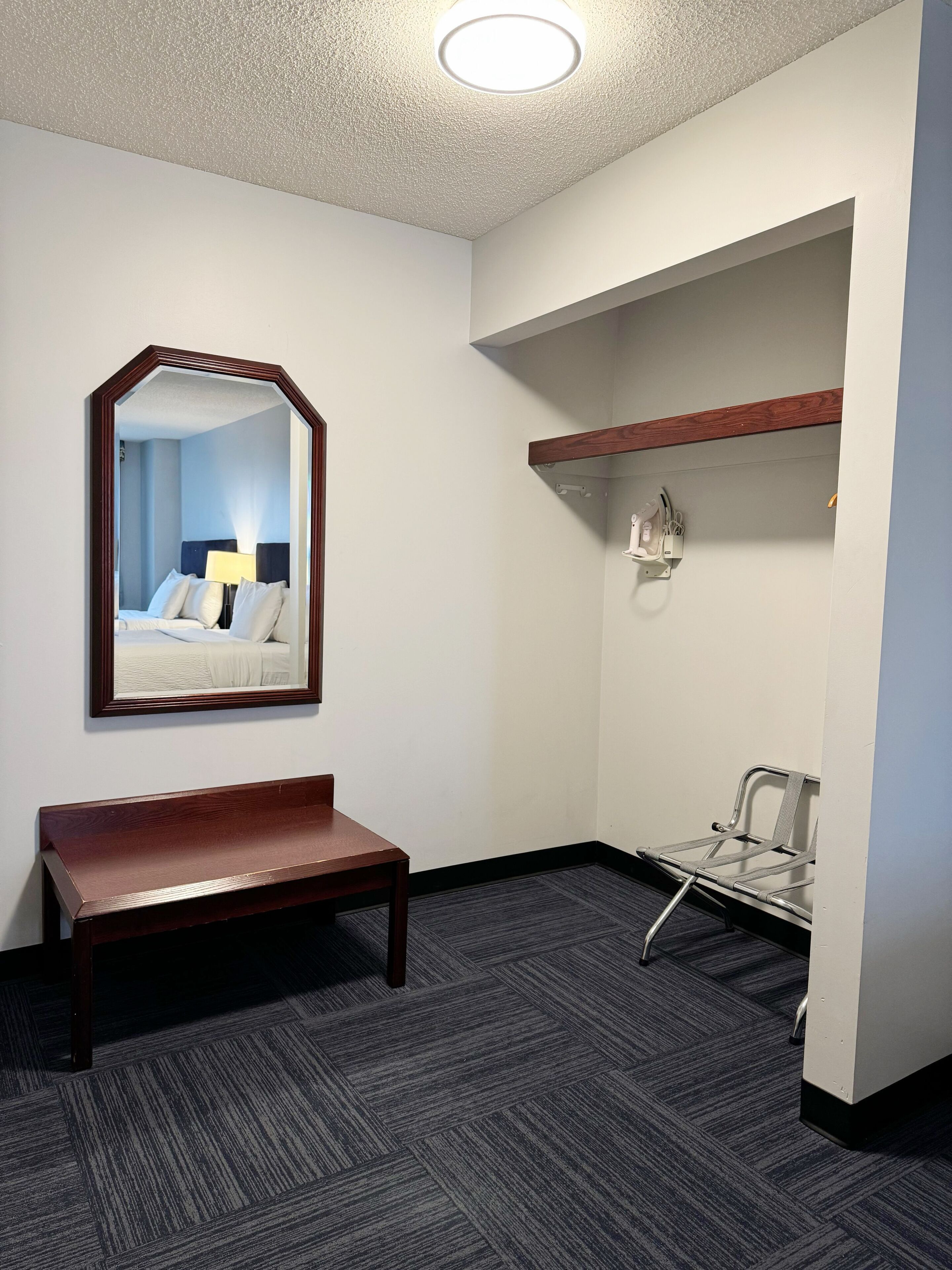 Ein Hotelzimmer mit einer Holzbank, einem Spiegel, einem Bett mit weißer Bettwäsche, einem kleinen Schreibtisch und einem Sessel.