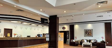 A hotel reception area with a reception desk, wall clocks, a large pillar, a sofa set, and a framed artwork on the wall.