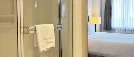 Habitación de hotel con cabina de ducha de vidrio, cama con cubrecama blanco, mesita de noche con lámpara y ventana con cortinas.