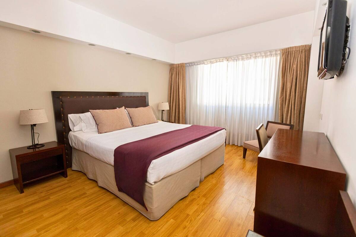 Une chambre d’hôtel avec un grand lit, une table de chevet en bois, un bureau et un téléviseur fixé au mur.