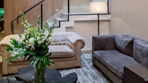 Un salon avec un fauteuil beige, un canapé gris, un vase de fleurs et un escalier en arrière-plan.