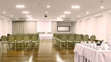 Un salón de conferencias con filas de sillas, una mesa dispuesta con tazas y platos, y una pantalla de proyector al frente.