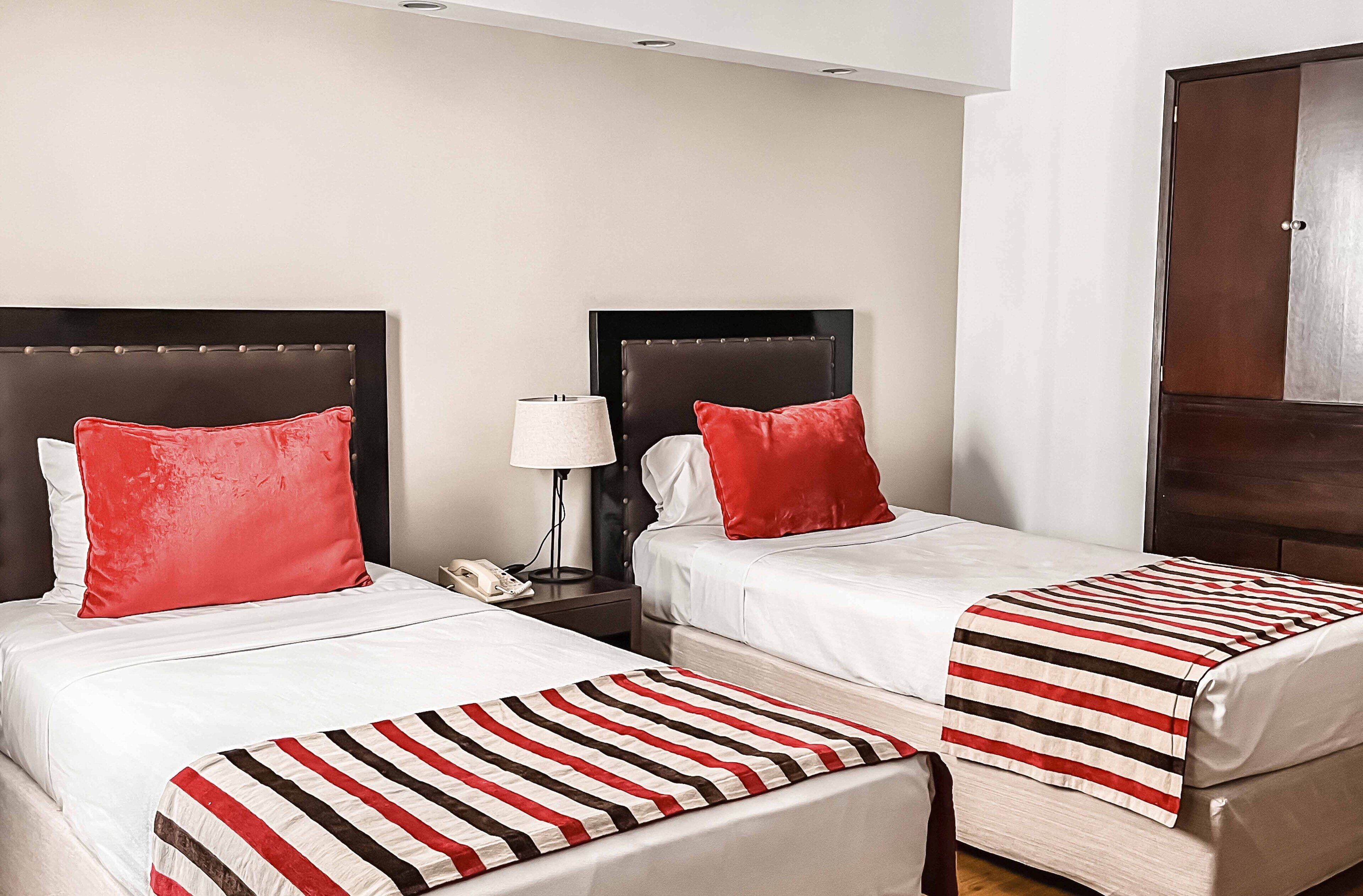 Two beds with red pillows, a bedside table with a lamp, and a striped blanket.