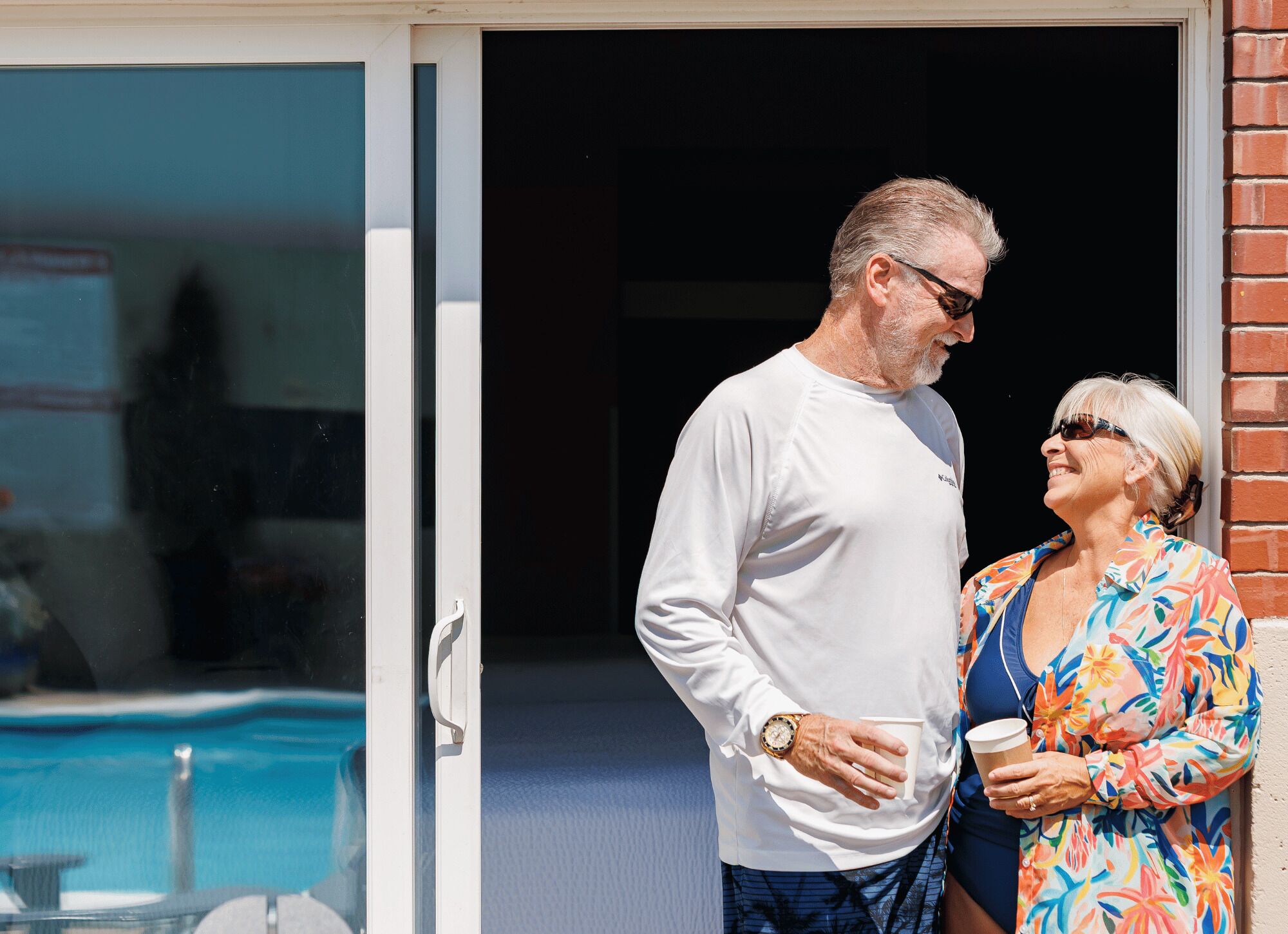 Un hombre y una mujer están de pie frente a un edificio, con una piscina visible a través de un ventanal.