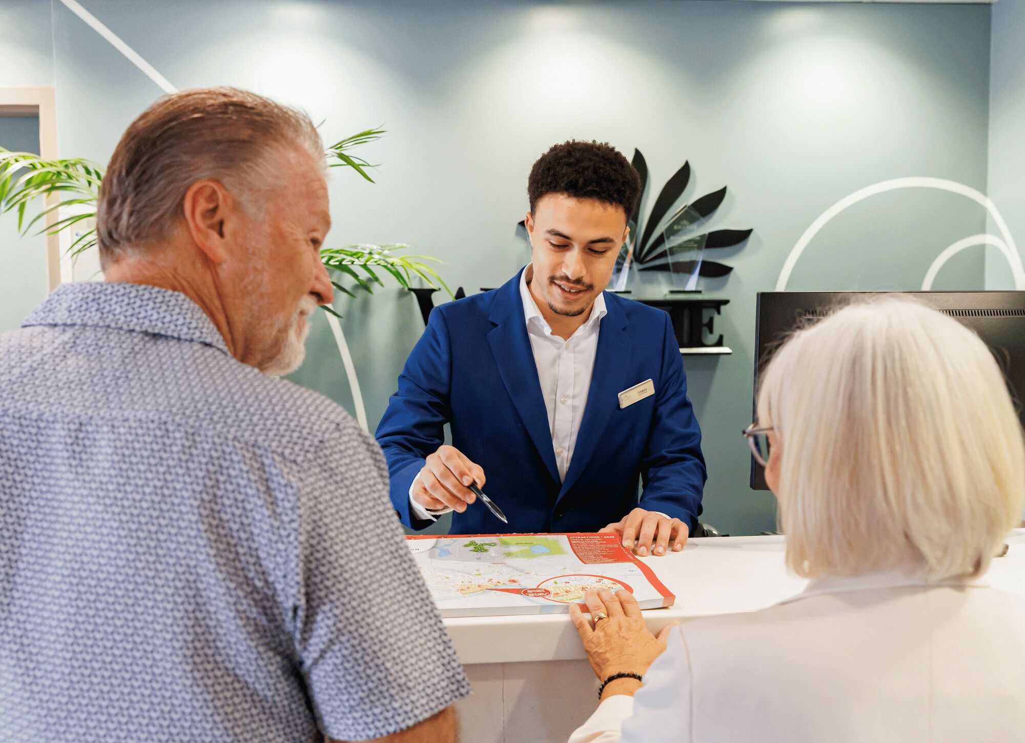 Une personne vêtue d’un costume bleu est assise à une table, une carte devant elle, et interagit avec deux autres personnes.