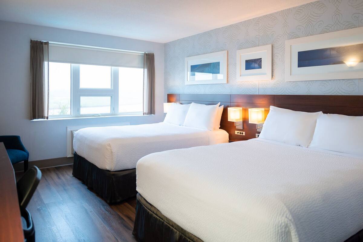 Quarto de hotel com duas camas, uma escrivaninha, uma cadeira e uma janela com cortinas.