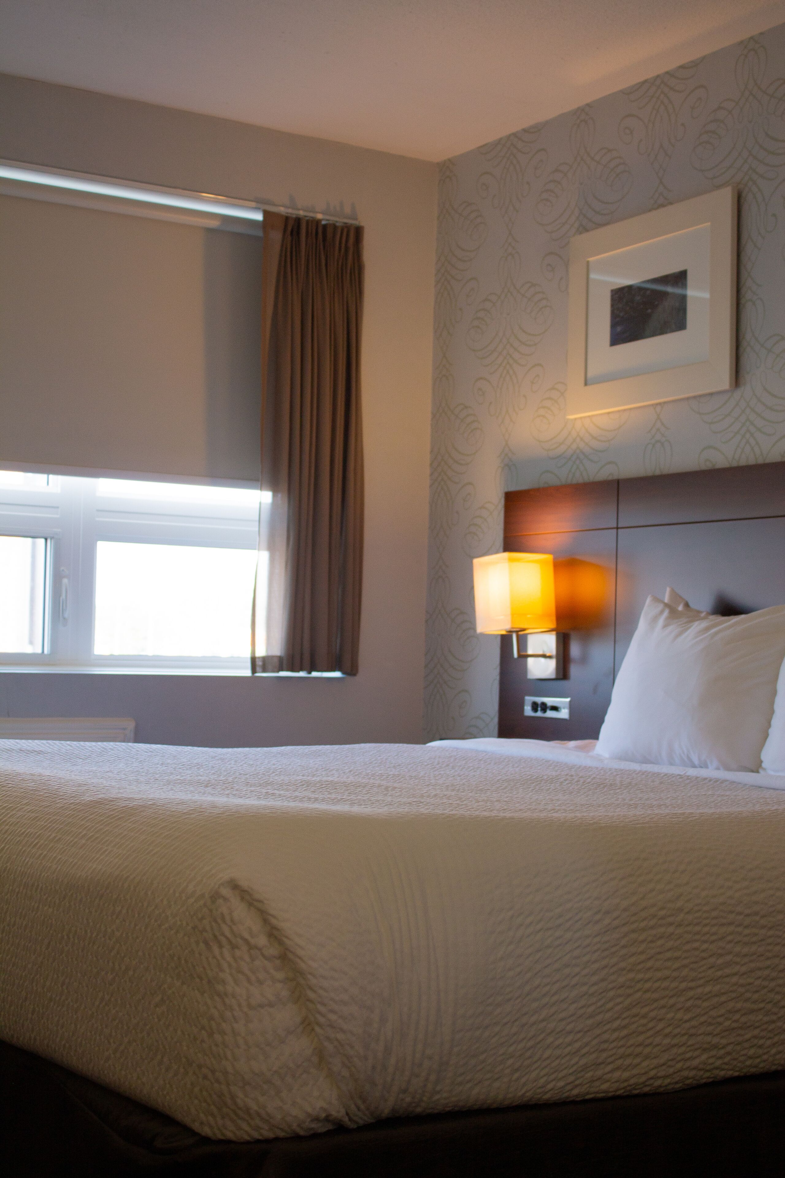 A hotel room with a large bed, a window with curtains, a wall-mounted lamp, and a framed picture.