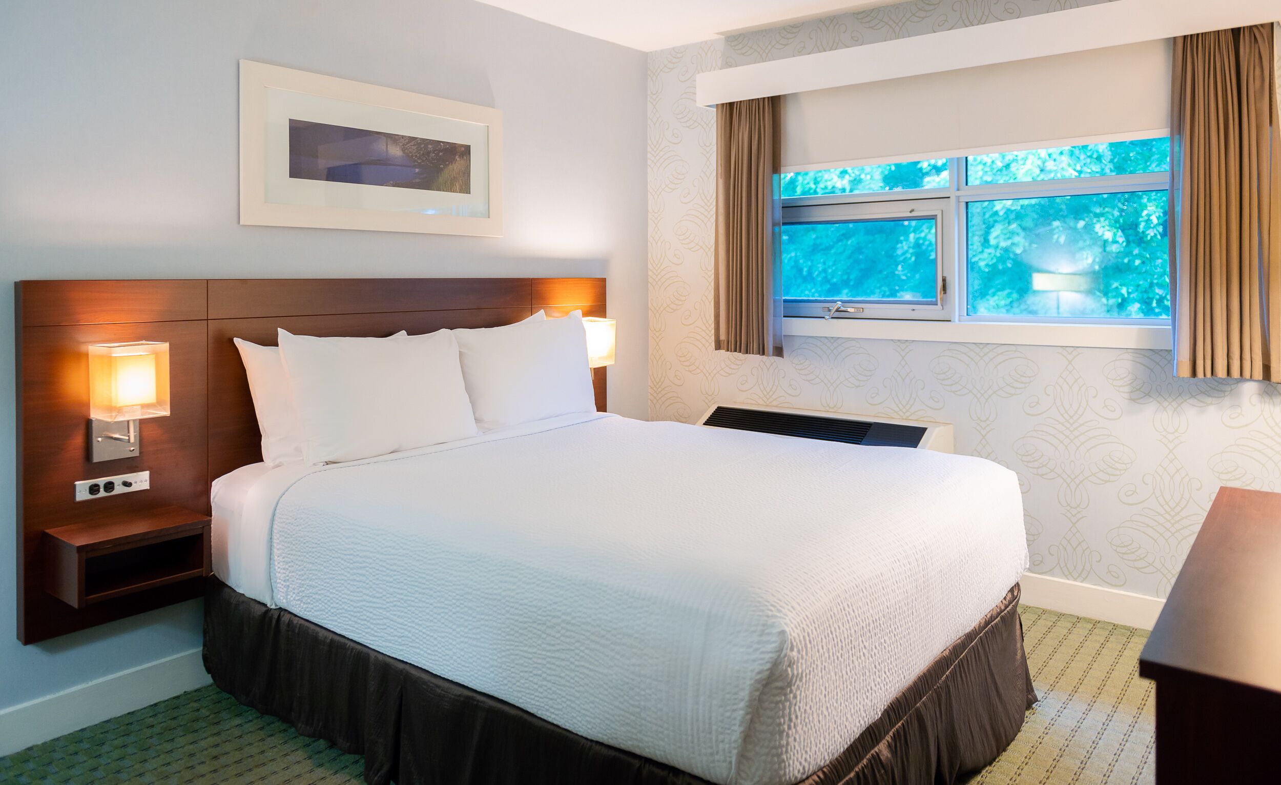 A hotel room with a large bed, two bedside lamps, a framed picture above the bed, a window with curtains, and a patterned wall.