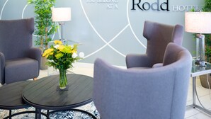 Um hall de hotel moderno com duas poltronas cinzas, uma mesa redonda e um vaso de flores amarelas. Uma placa na parede diz "Rodd Hotel" e "Você se sente em casa no Rodd Hotel".