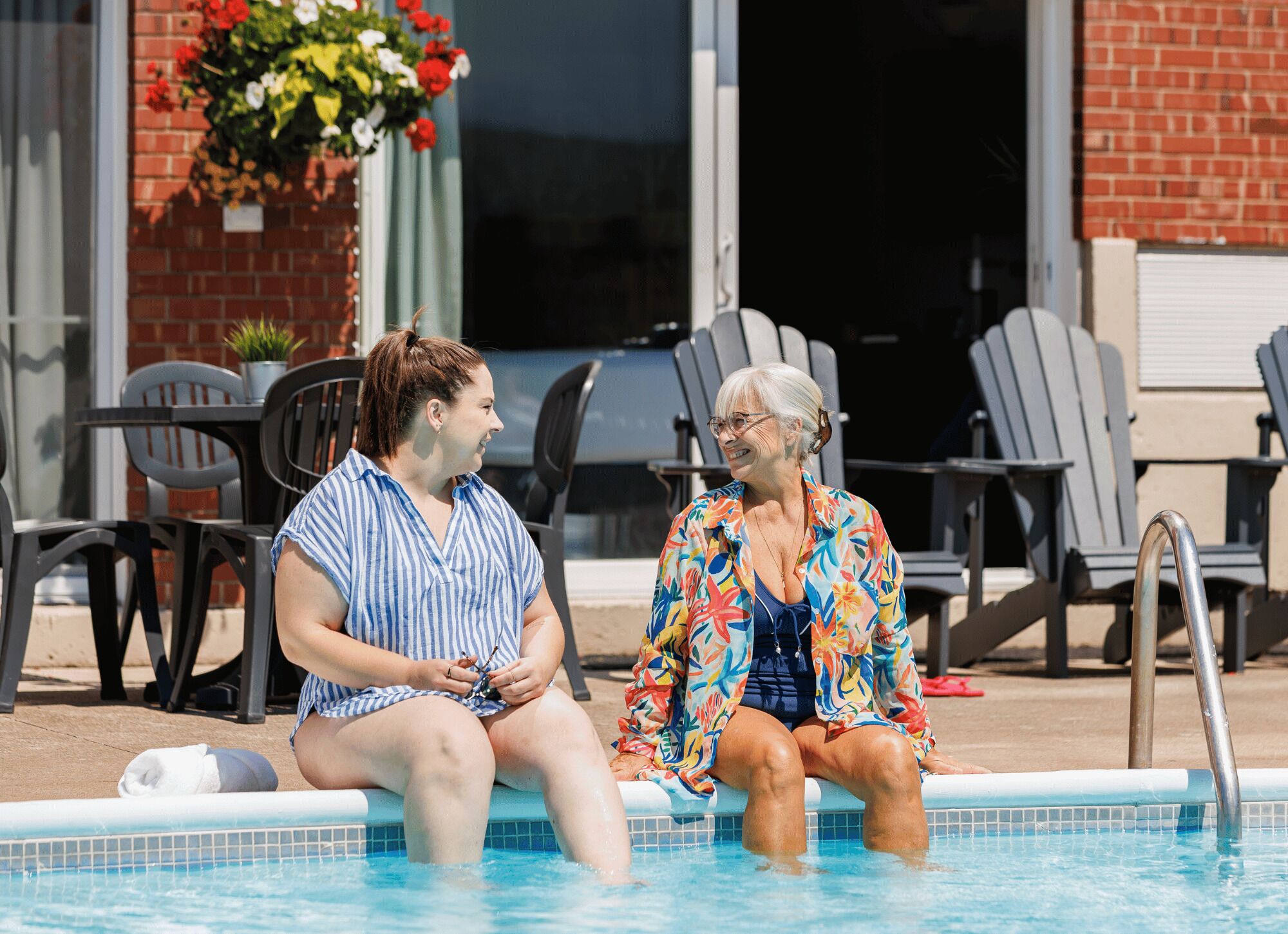 Two individuals seated by a poolside with lounge chairs and a brick wall in the background.