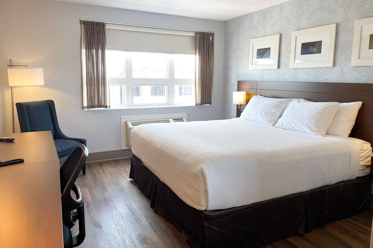 Quarto de hotel com cama, escrivaninha, cadeira, janela com cortinas e quadros nas paredes.