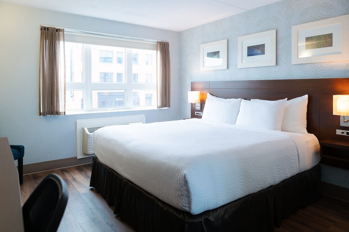 Quarto de hotel com uma cama grande, duas luminárias de cabeceira, uma janela com cortinas e quadros nas paredes.