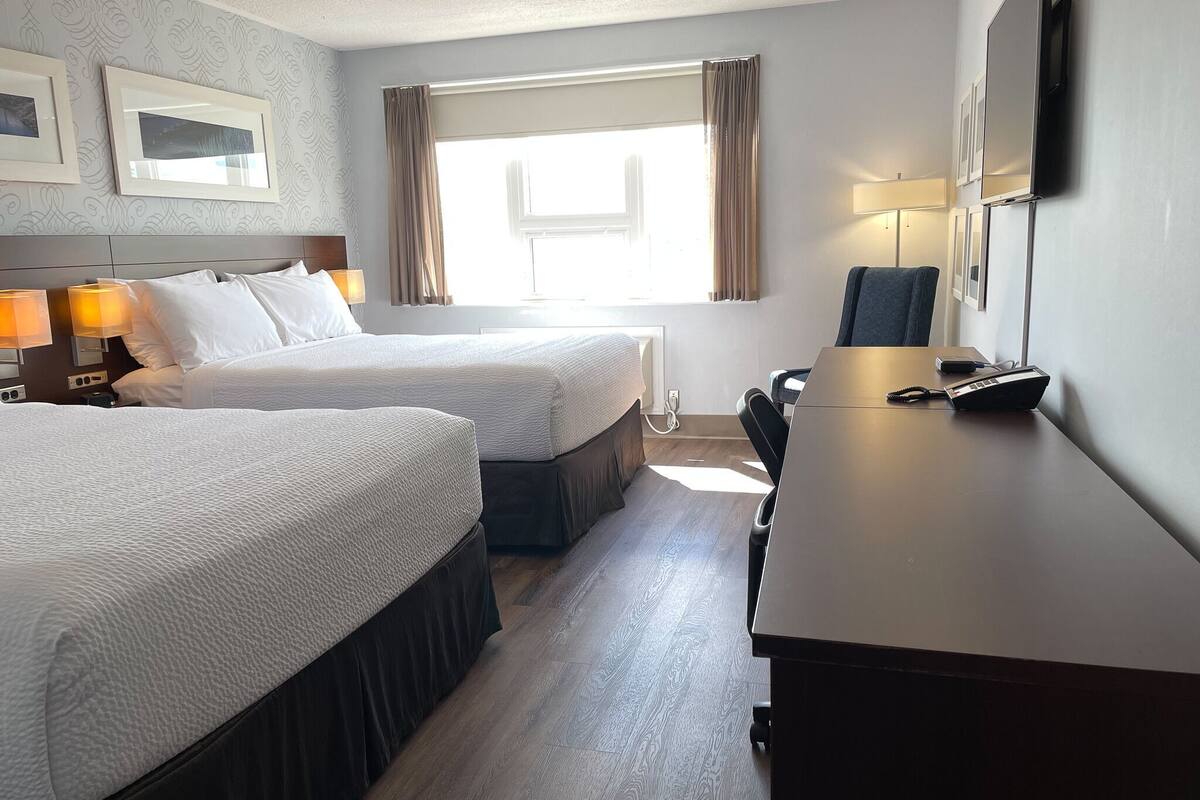 Quarto de hotel com duas camas, uma escrivaninha com telefone e janela com cortinas.