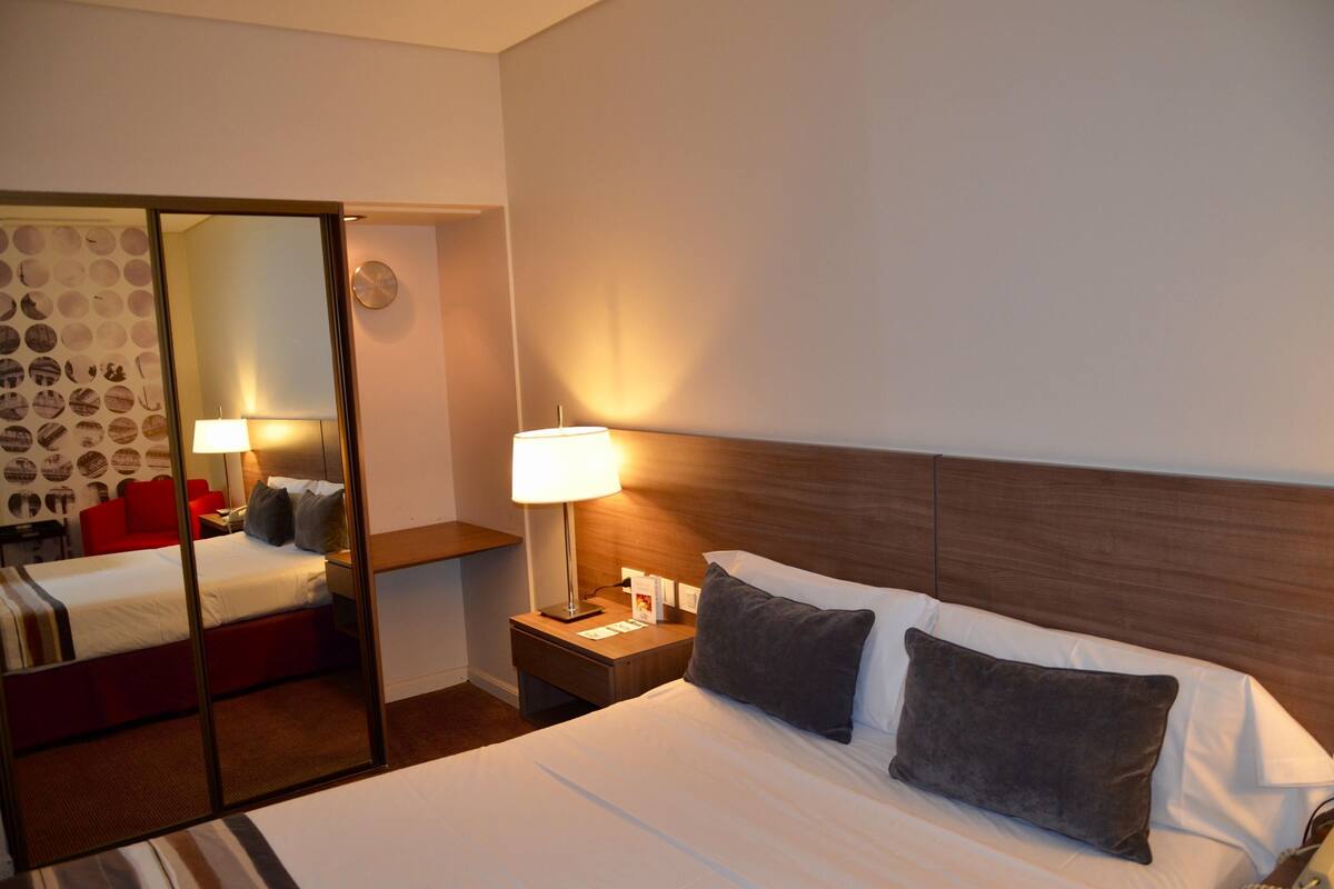 Una camera d'albergo con un letto, un comodino, una lampada, uno specchio e una sedia rossa.