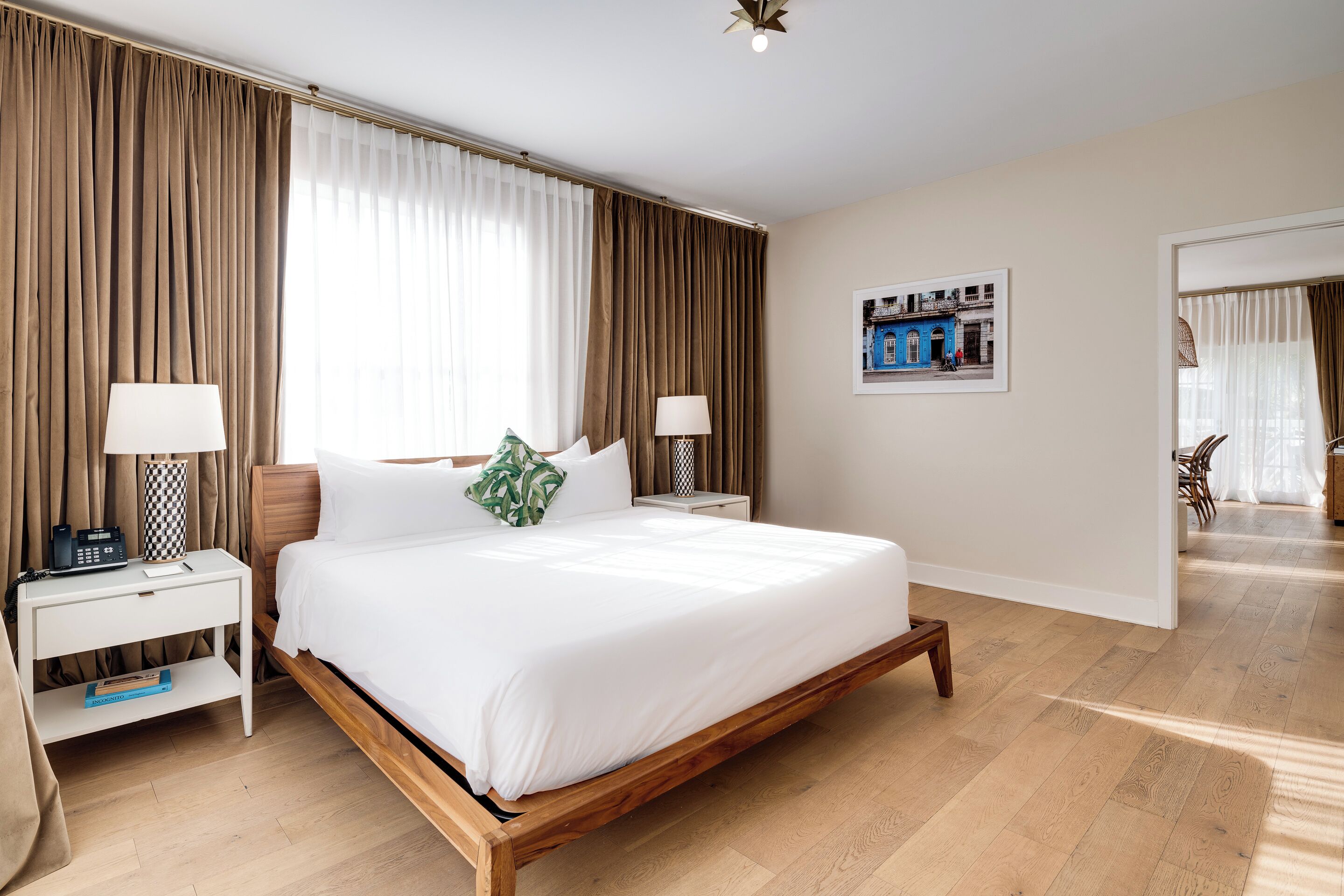 A hotel room with a wooden bed, white bedding, a nightstand with a lamp, a small table with a book, and a framed picture on the wall.