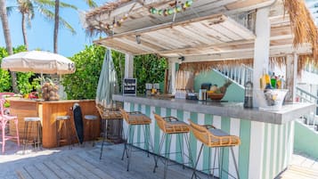 A beachside bar with a wooden deck, striped bar counter, and wicker bar stools.