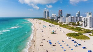 A beach with a row of blue beach chairs and umbrellas, a lifeguard tower, and tall buildings in the background. - The Savoy Hotel & Beach Club (Miami Beach)