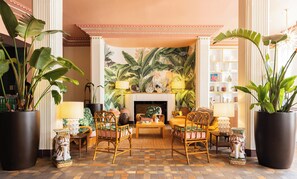 A spacious living room with a fireplace, large potted plants, wicker furniture, and a tropical mural on the wall. - The Savoy Hotel & Beach Club (Miami Beach)