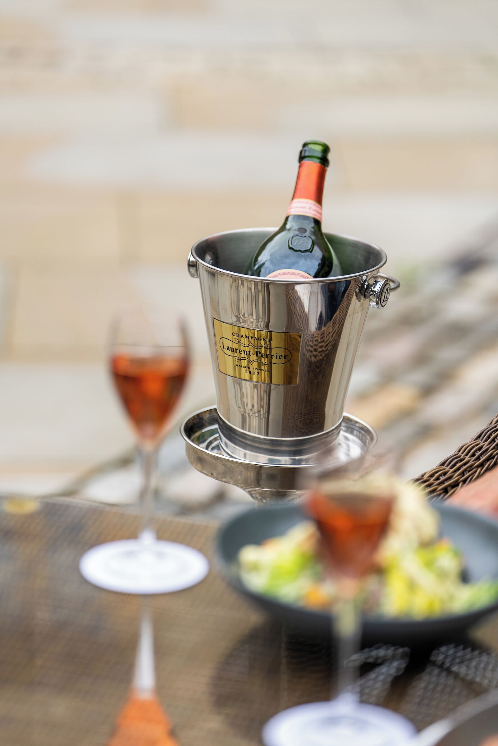 A bottle of champagne in a metal bucket with a label, a glass of wine, and a plate of food on a table.