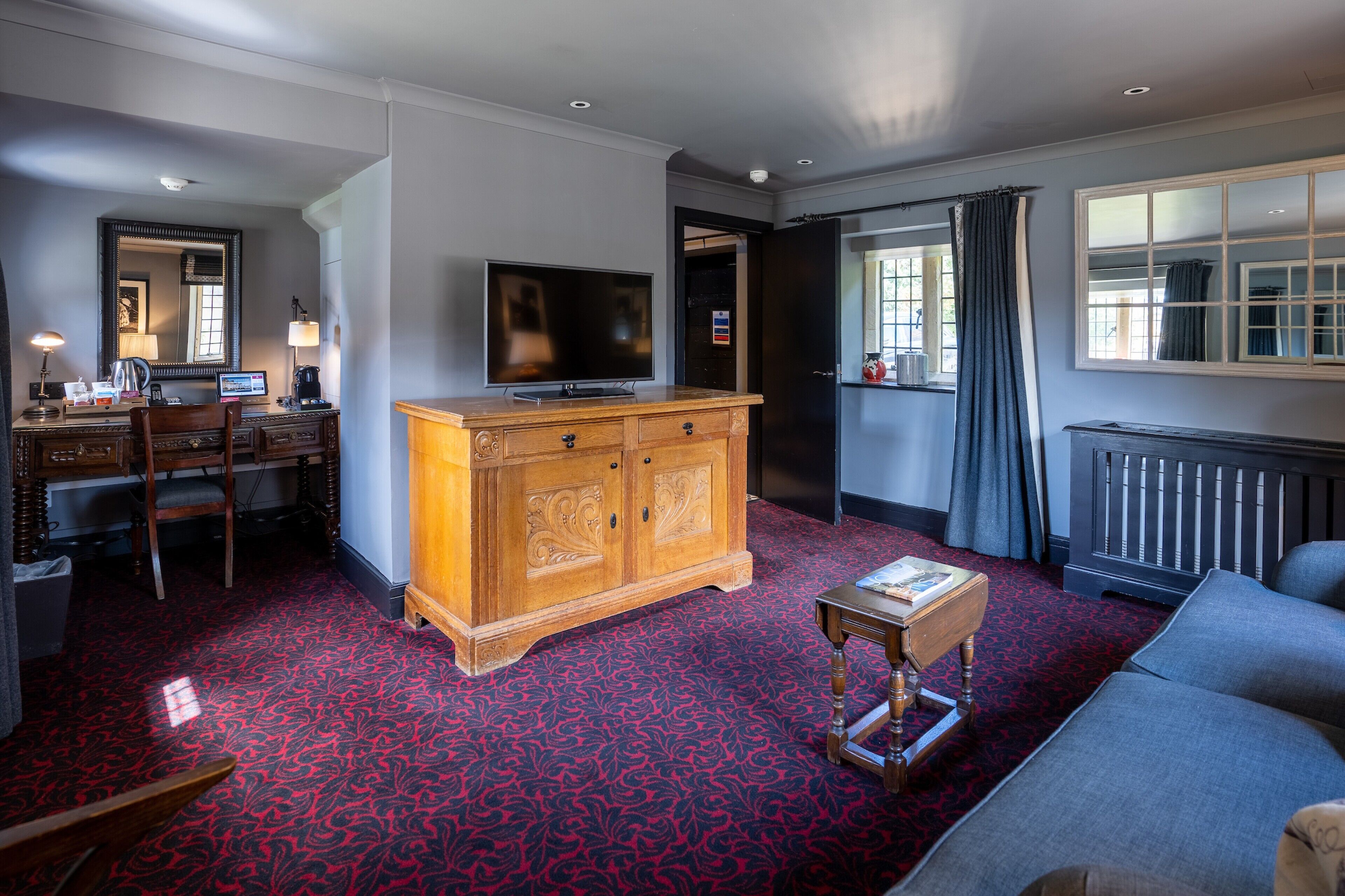 A hotel room with a red patterned carpet, a wooden cabinet, a television, a sofa, and a desk with a computer.
