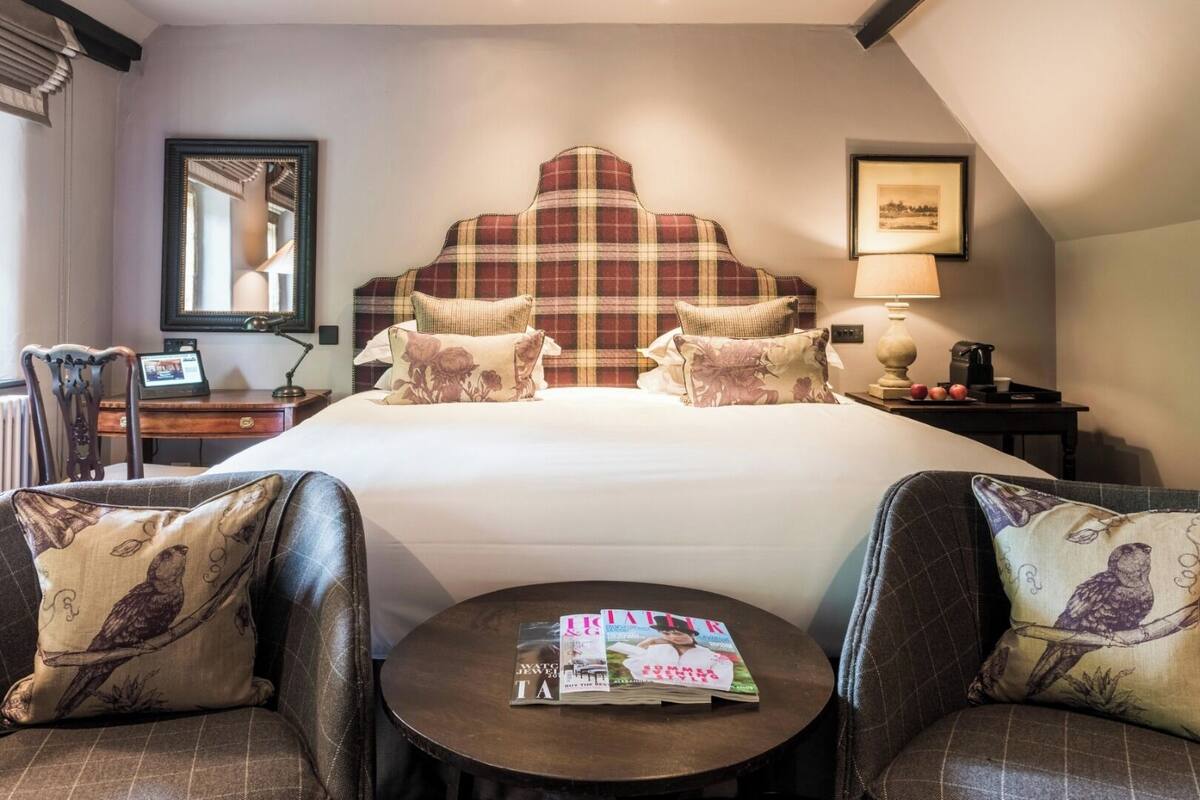 A hotel room with a large bed, two armchairs, a small round table with magazines, a desk with a computer, and a framed picture on the wall.