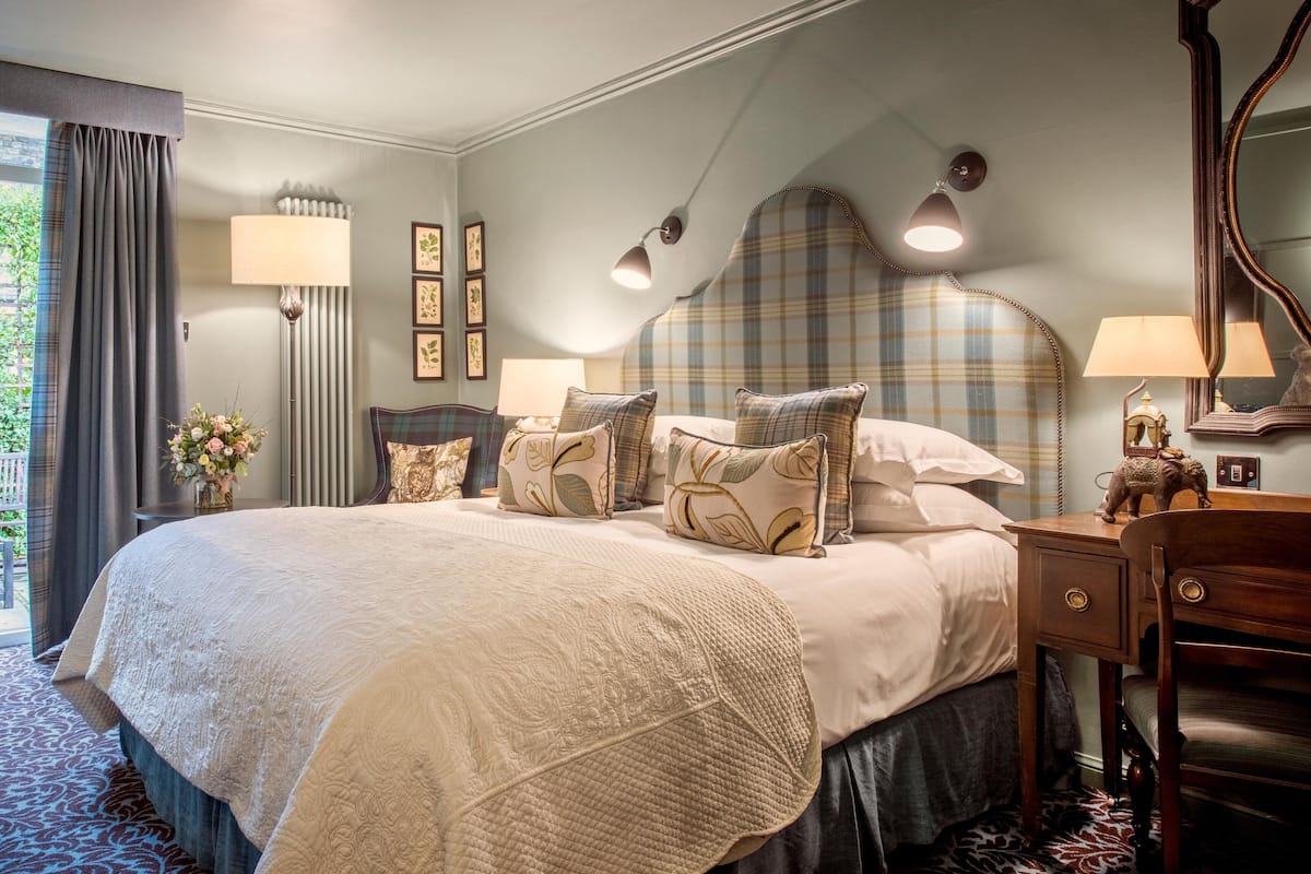 A well-appointed bedroom with a large bed, multiple pillows, a bedside table with a lamp, and a view of greenery through a window.