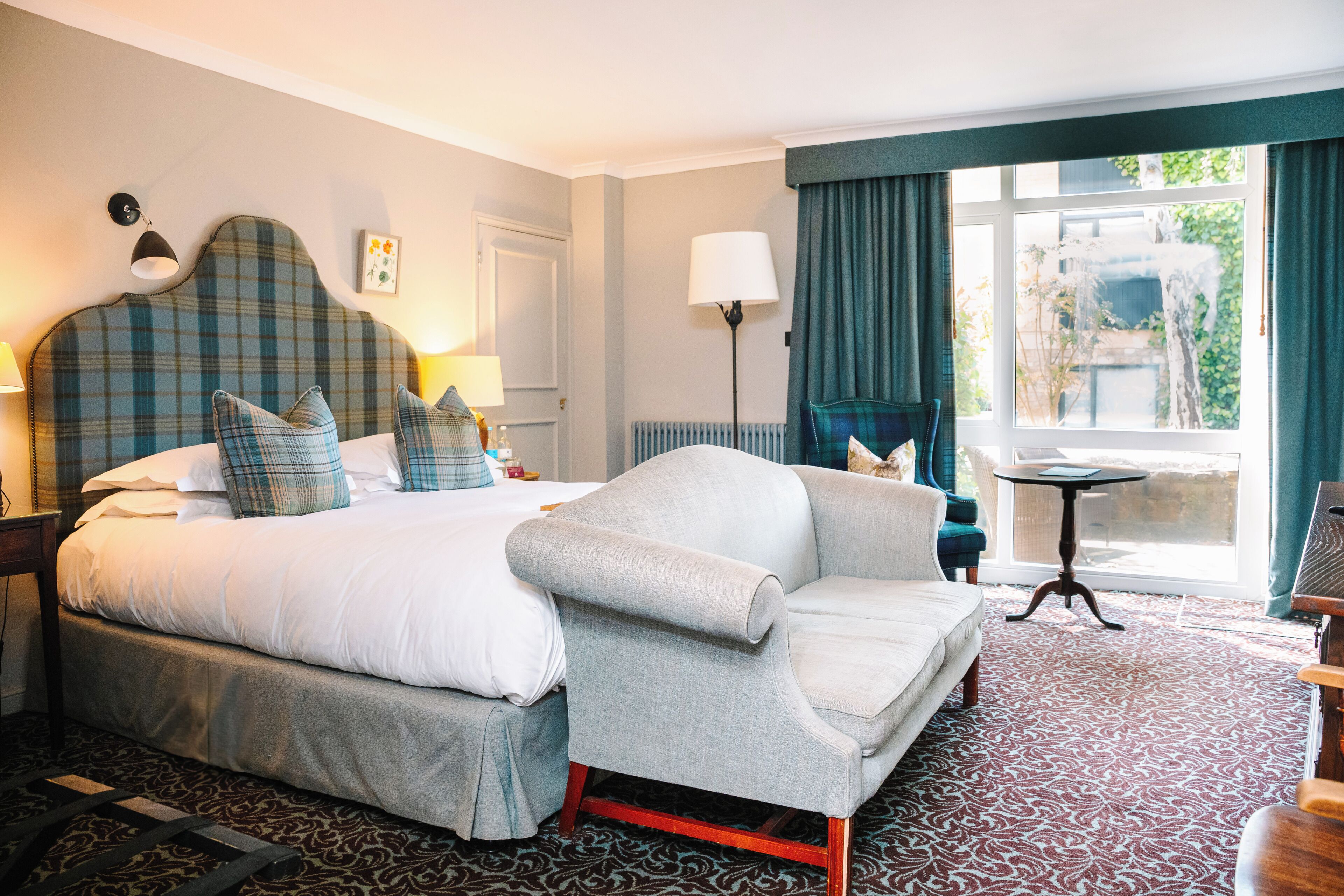 A hotel room with a large bed, a grey armchair, a small table, and a view of greenery outside.
