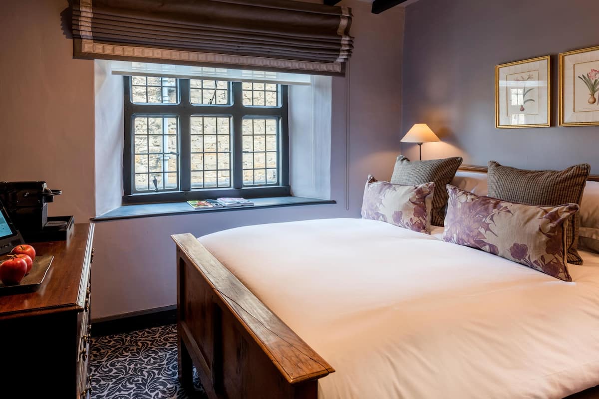 A bedroom with a large bed, a window with blinds, a desk with a laptop and a basket of tomatoes, and two framed botanical prints on the wall.