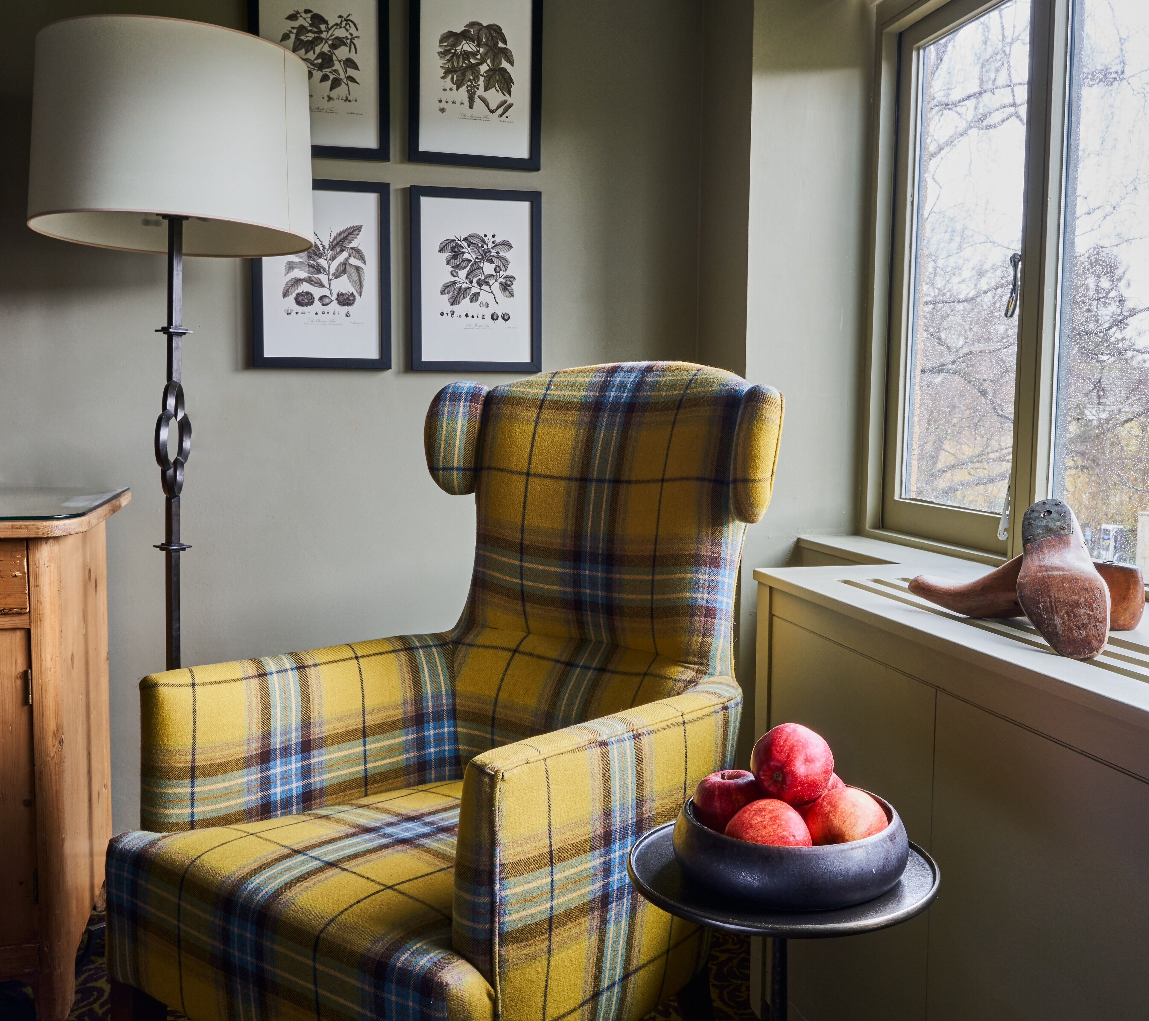 A plaid armchair by a window with a bowl of apples and a lamp.
