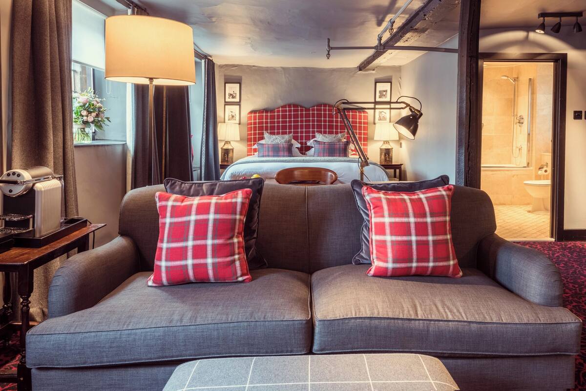 A modern hotel room with a large bed, a sofa with plaid cushions, a desk with a lamp, and a view of the bathroom.