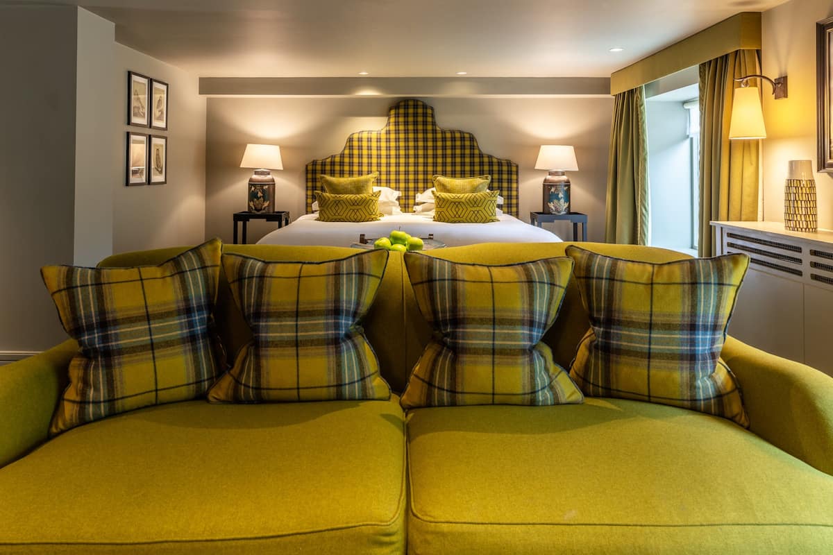 A hotel room with a large bed, two bedside tables with lamps, a green sofa with plaid cushions, and a headboard with a plaid pattern.