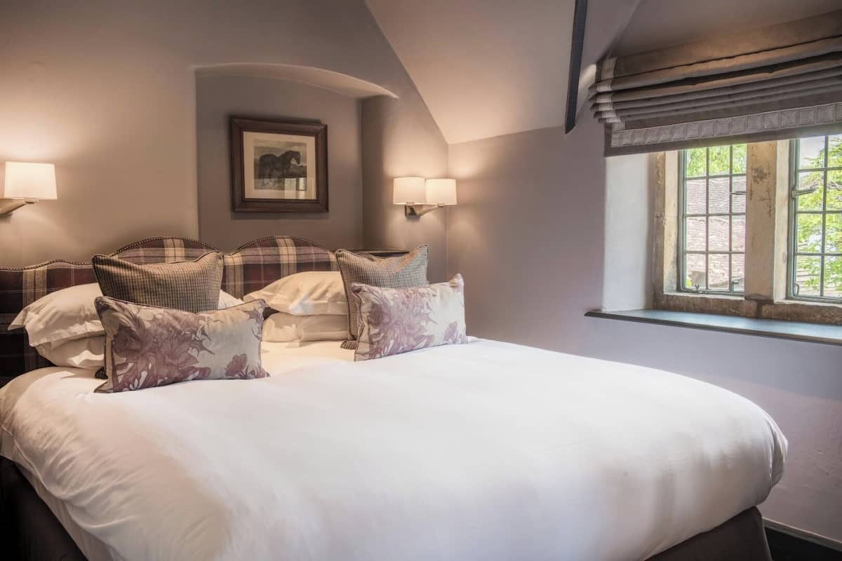 A neatly made bed with a headboard, two bedside lamps, a framed picture on the wall, and a window with a view of greenery.