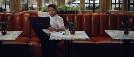 A man sitting on a leather bench in a restaurant with a menu titled "GRILL" and two small tables with vases.