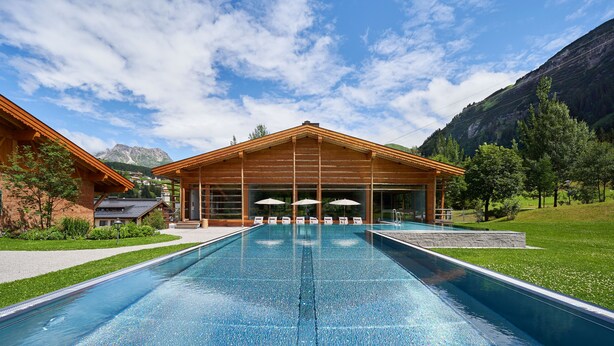 Zona de piscina con un edificio de madera, mobiliario exterior y montañas al fondo.