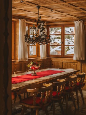 Comedor de madera con una mesa dispuesta para seis comensales, una araña y vistas de casas nevadas a través de amplios ventanales.
