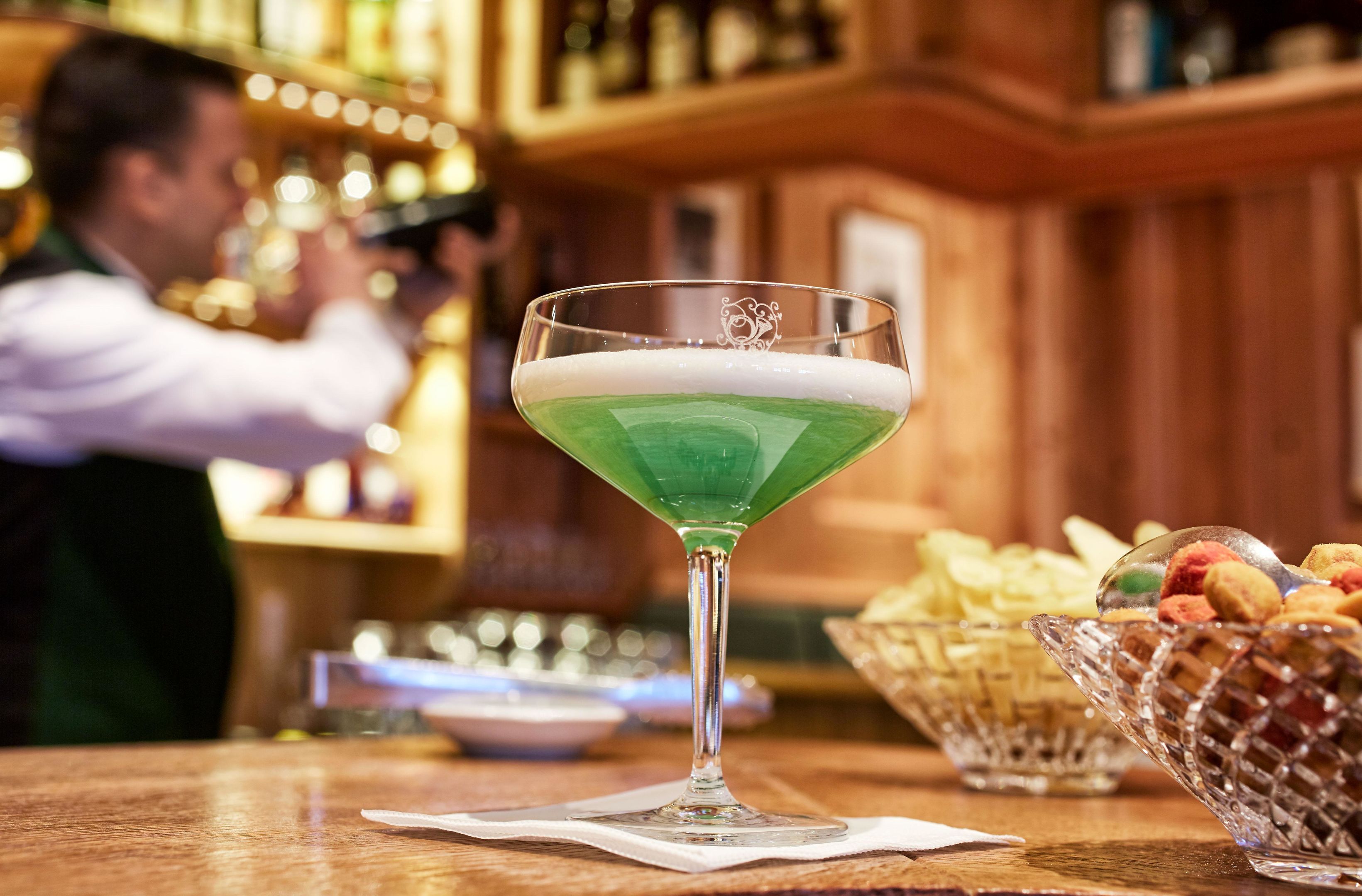 Un bar con una copa de martini sobre un posavasos, una cesta de aperitivos y un bartender desenfocado al fondo.