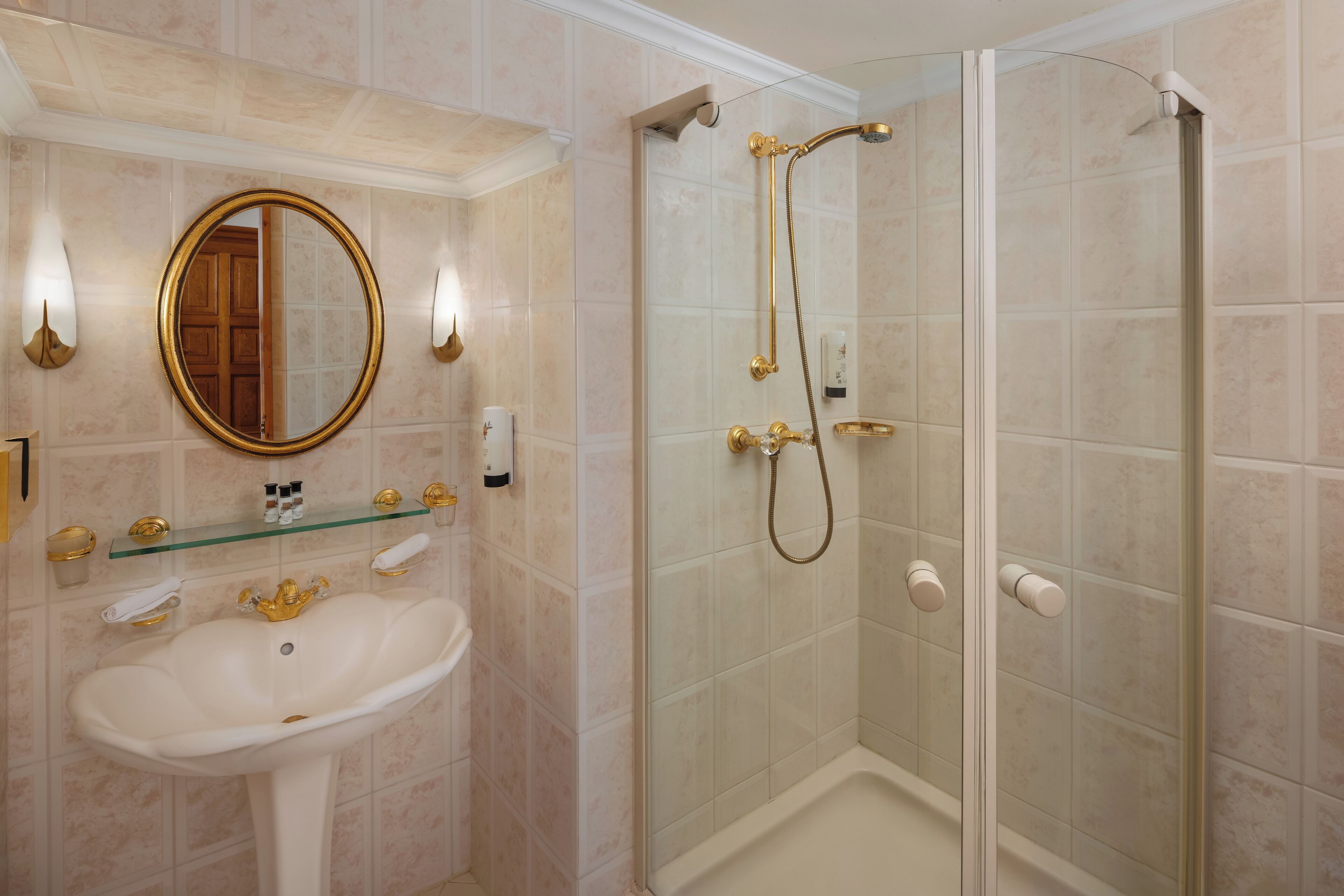 Un baño con lavamanos pedestal, espejo, ducha con mampara de vidrio y estante de vidrio con artículos de aseo.