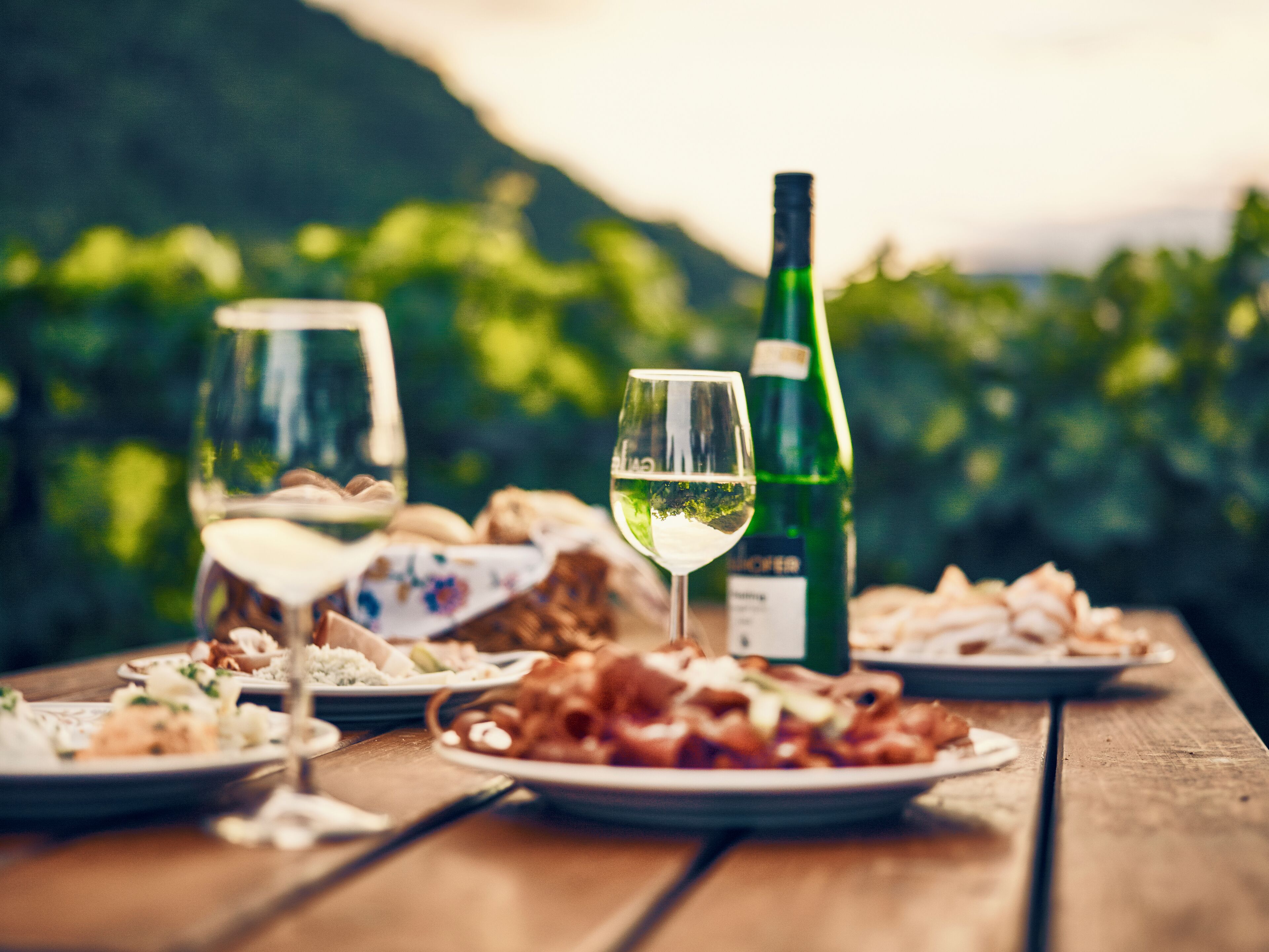 A wooden table with plates of food and wine glasses, set outdoors with a blurred green background.