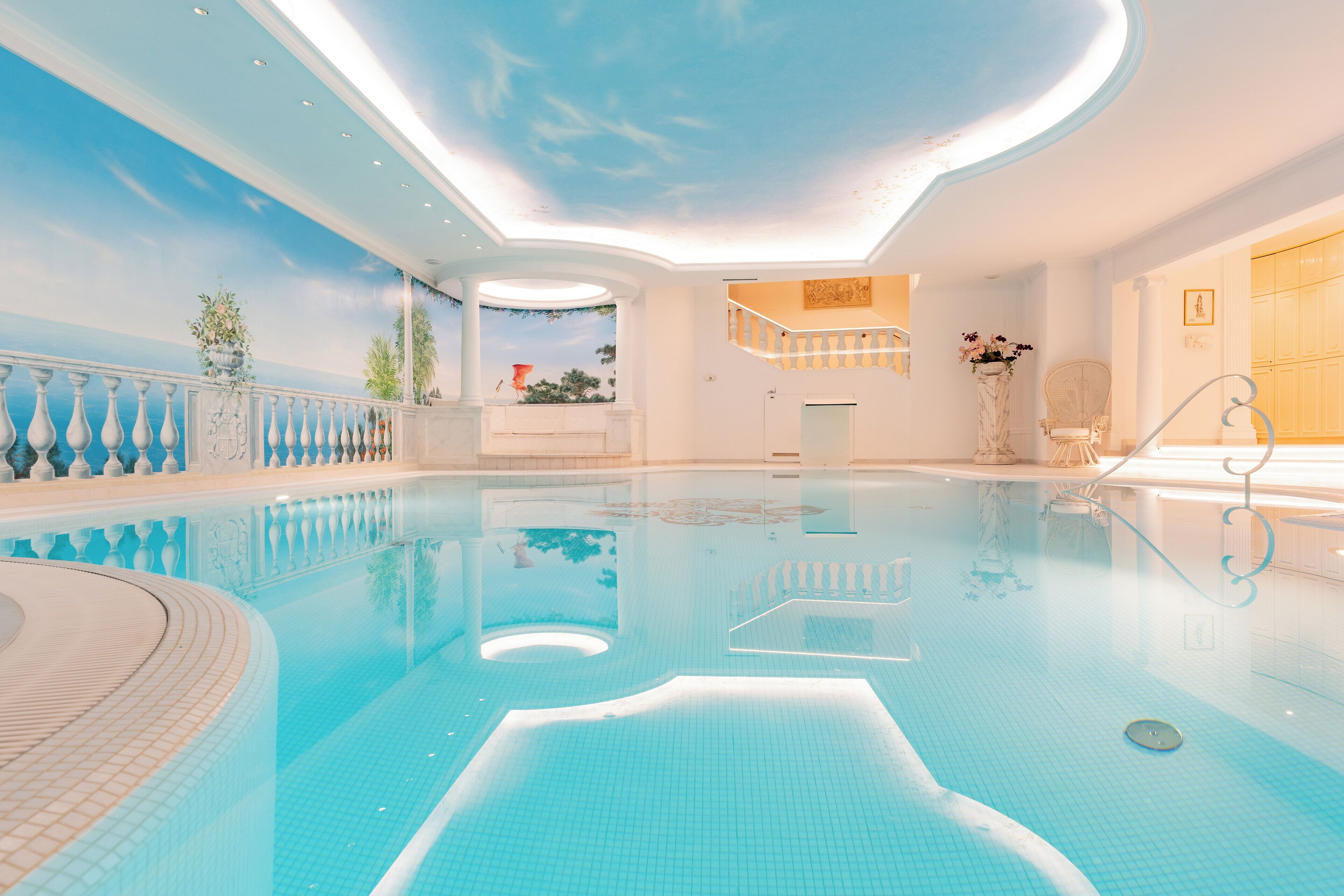Ein großer, überdachter Schwimmbereich mit Deckenfresko, einer eingelassenen Sitzecke und einer Treppe, die zu einer erhöhten Ebene führt.