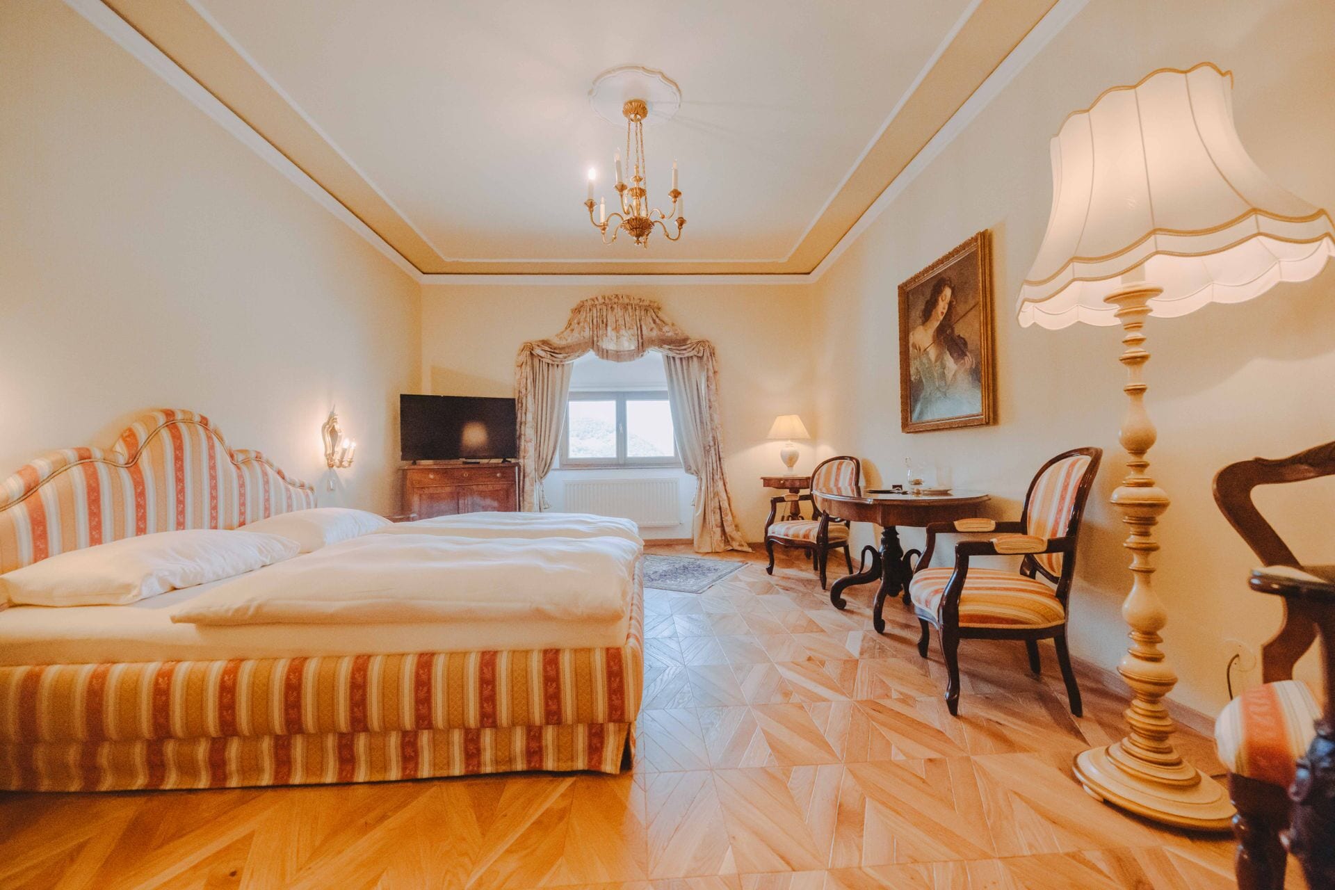 A hotel room with a large bed, a desk, two chairs, a television, and a chandelier.