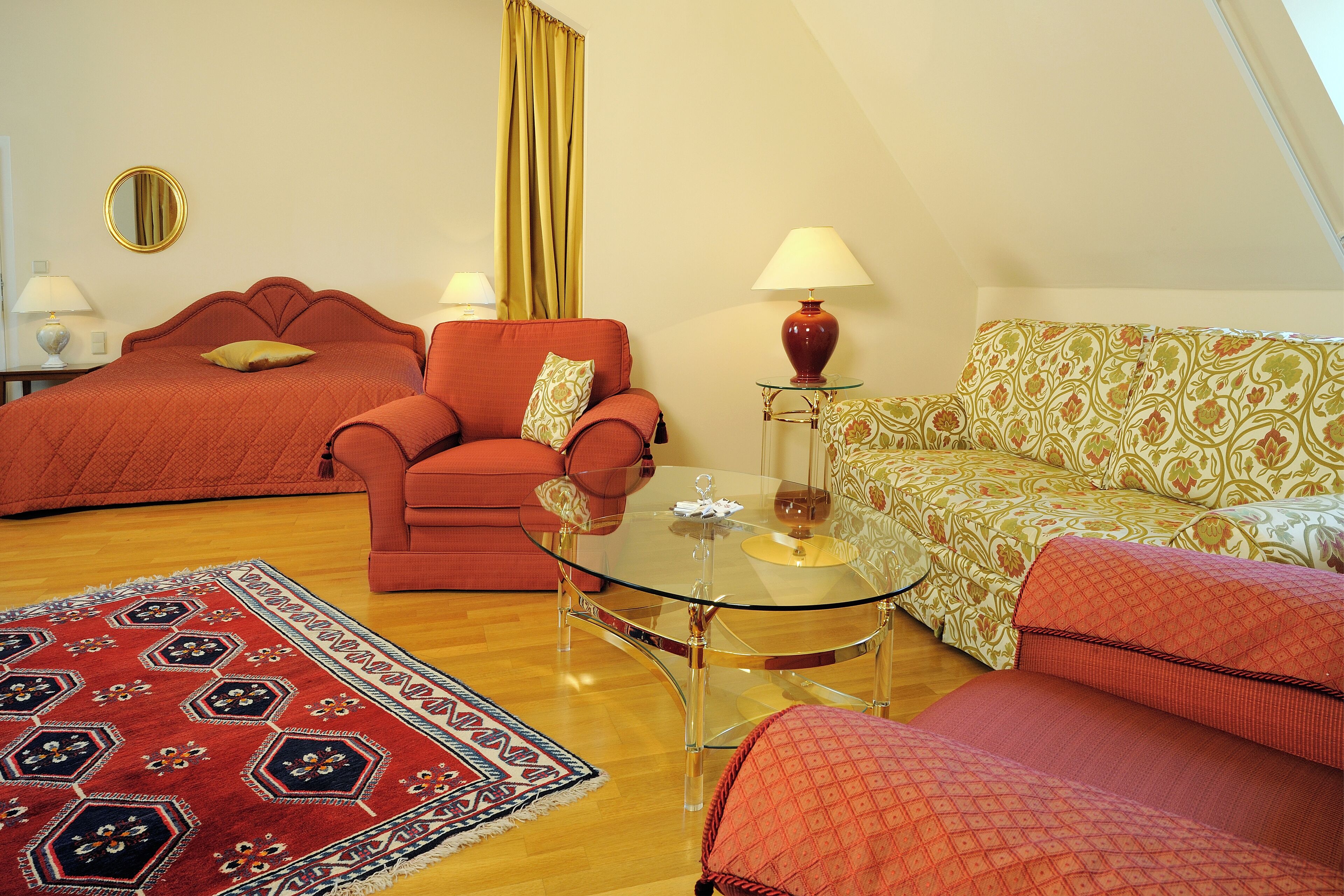 Una habitación acogedora con una cama, un sofá, una mesa de centro de vidrio y una alfombra estampada.
