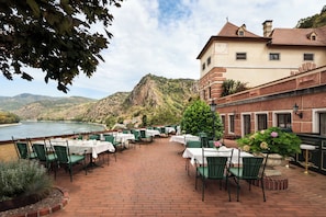 A riverside outdoor dining area with brick flooring, green chairs, and white tablecloths. - Hotel Schloss Durnstein (Durnstein)