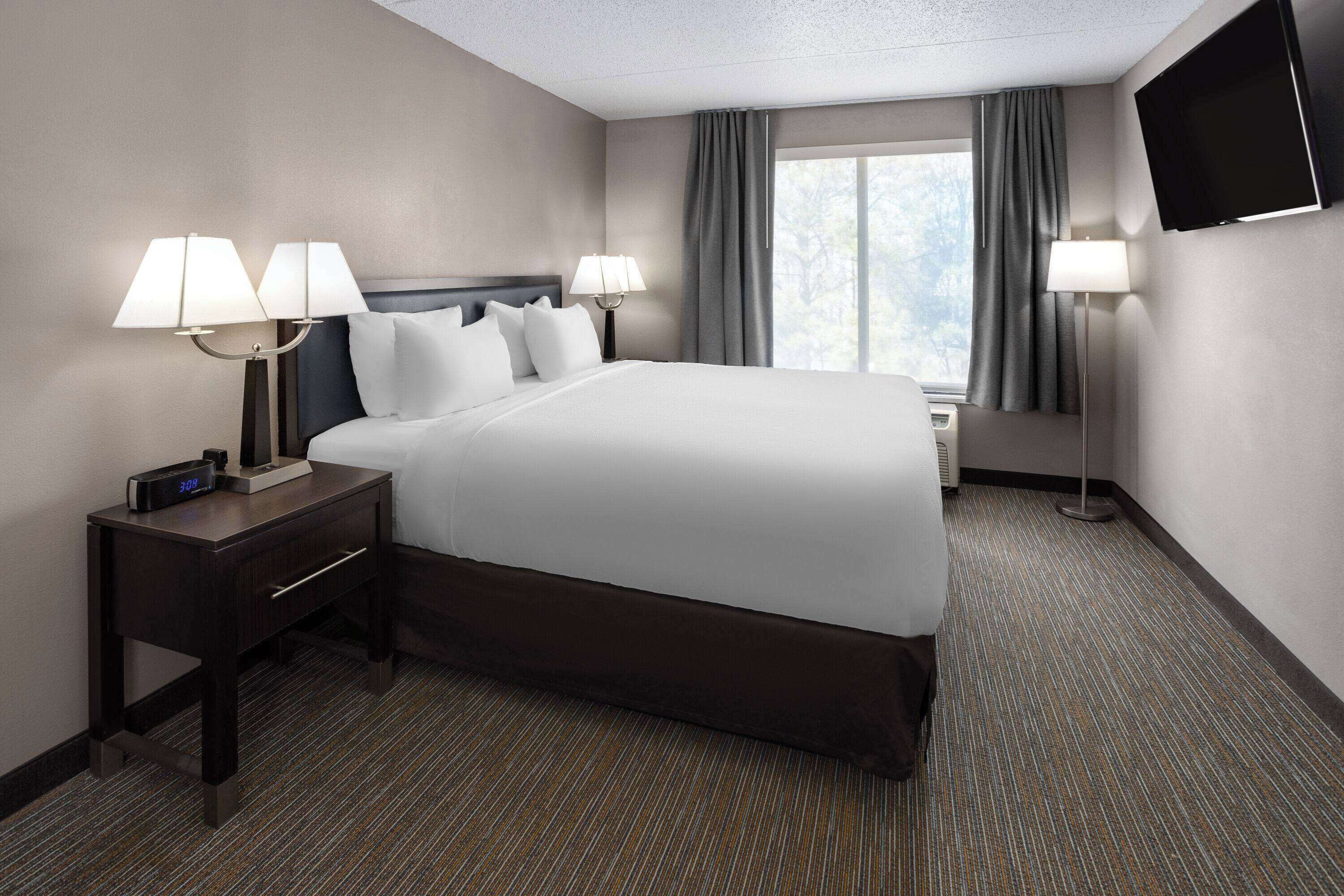 A hotel room with a large bed, two bedside tables with lamps, a flat-screen TV mounted on the wall, and a window with curtains.