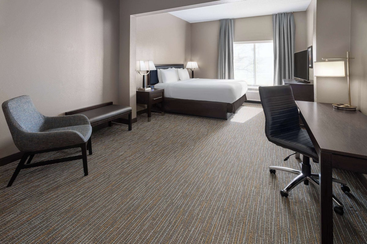 A hotel room with a bed, two chairs, a desk, a TV, and a lamp.