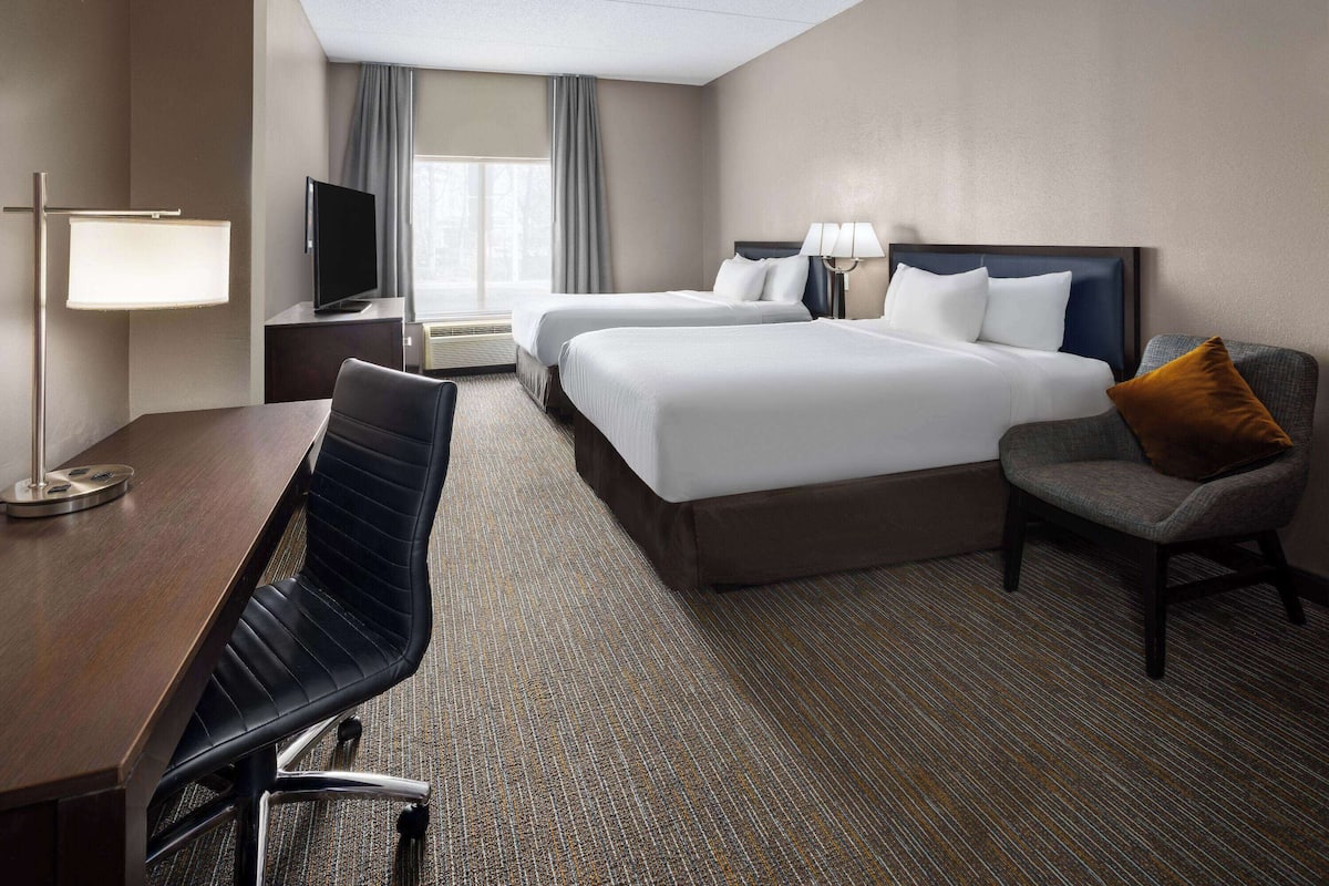 A hotel room with two beds, a desk, a chair, a TV, and a lamp.