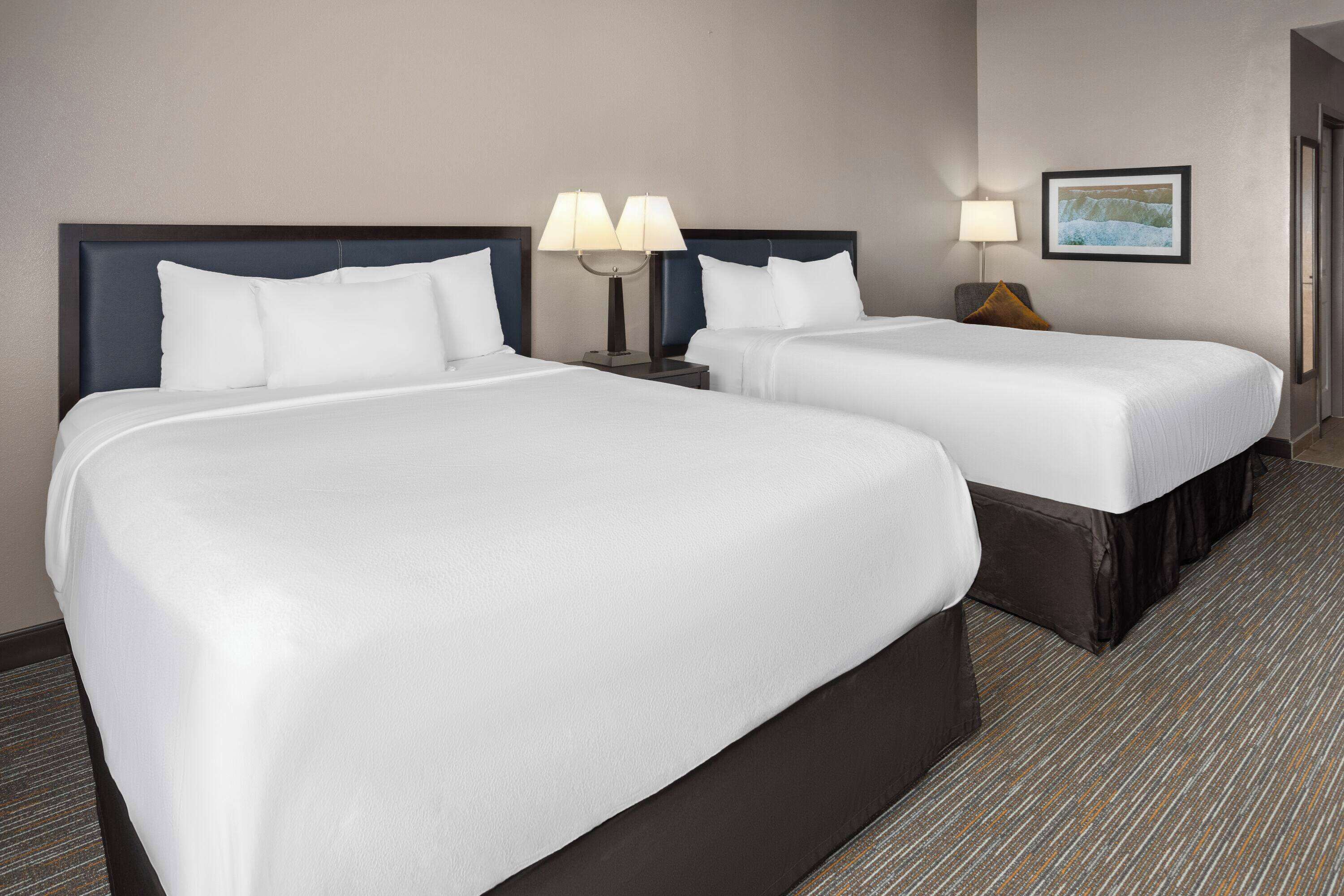 A hotel room with two beds, each with white linens and dark headboards. There are two bedside tables with lamps, and a framed picture on the wall.