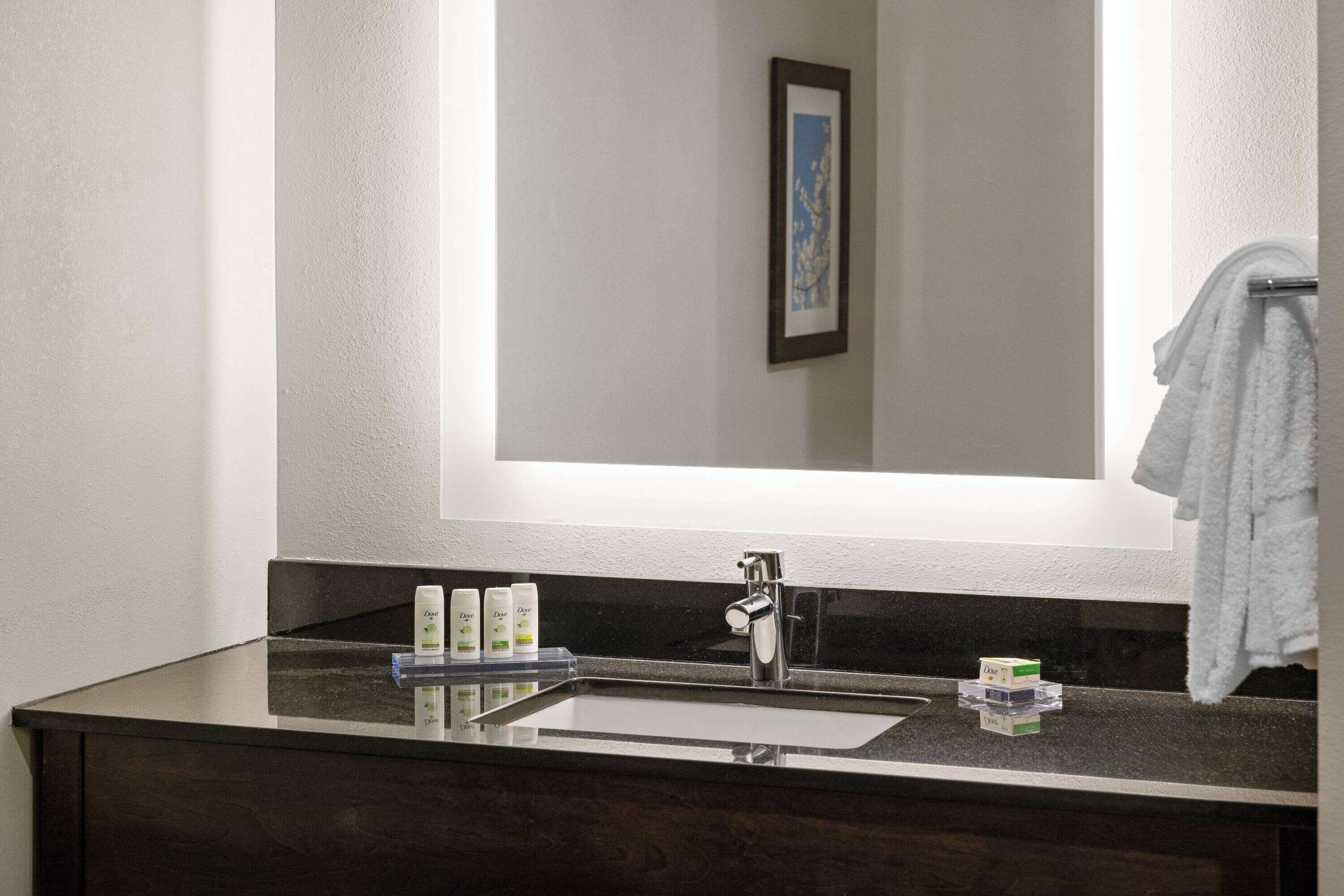 Une salle de bain avec un meuble-lavabo en bois foncé, un lavabo blanc et un miroir au-dessus. On y trouve trois bouteilles de savon Dove sur le meuble-lavabo.