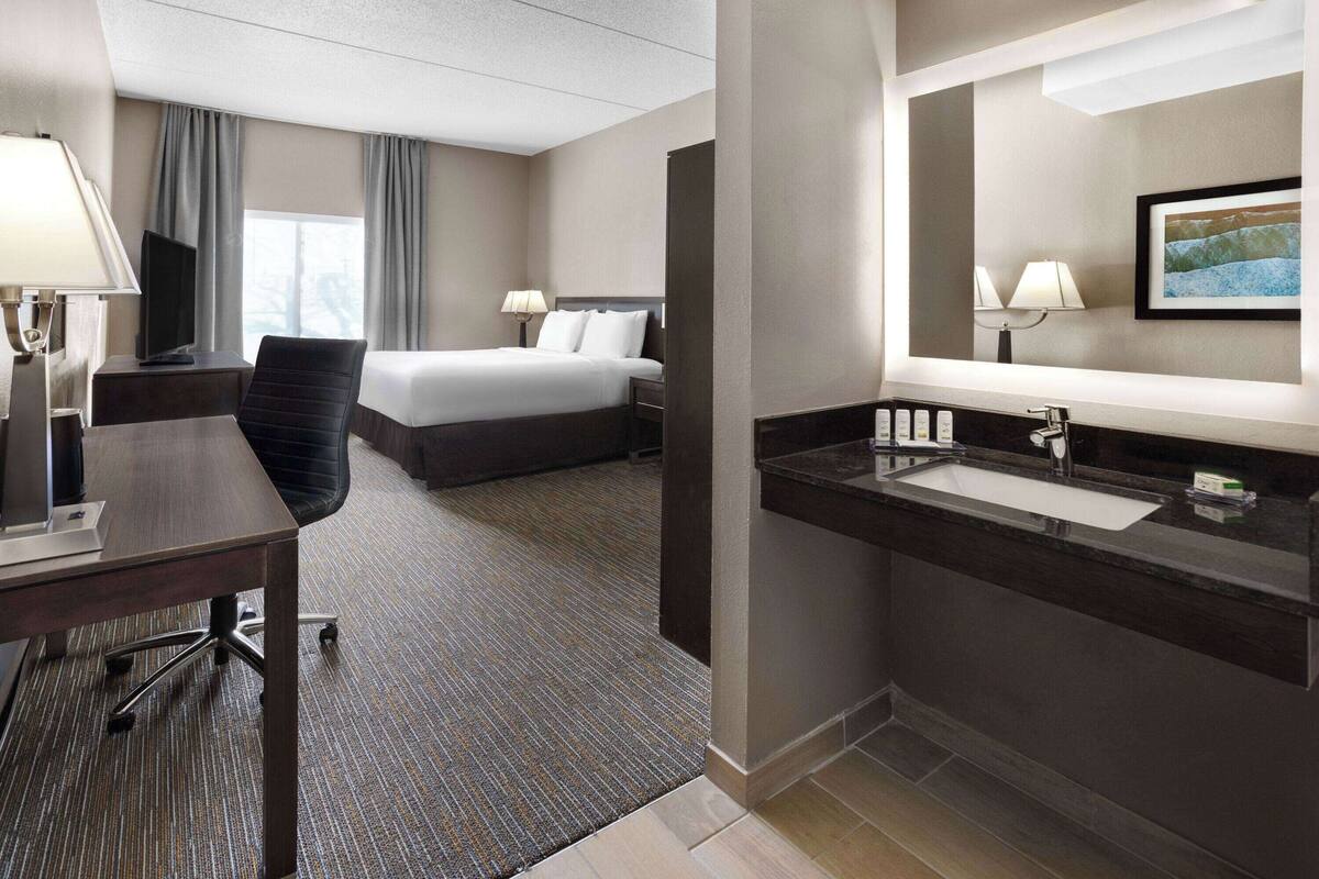 A hotel room with a bed, desk, chair, and a bathroom with a sink and mirror.