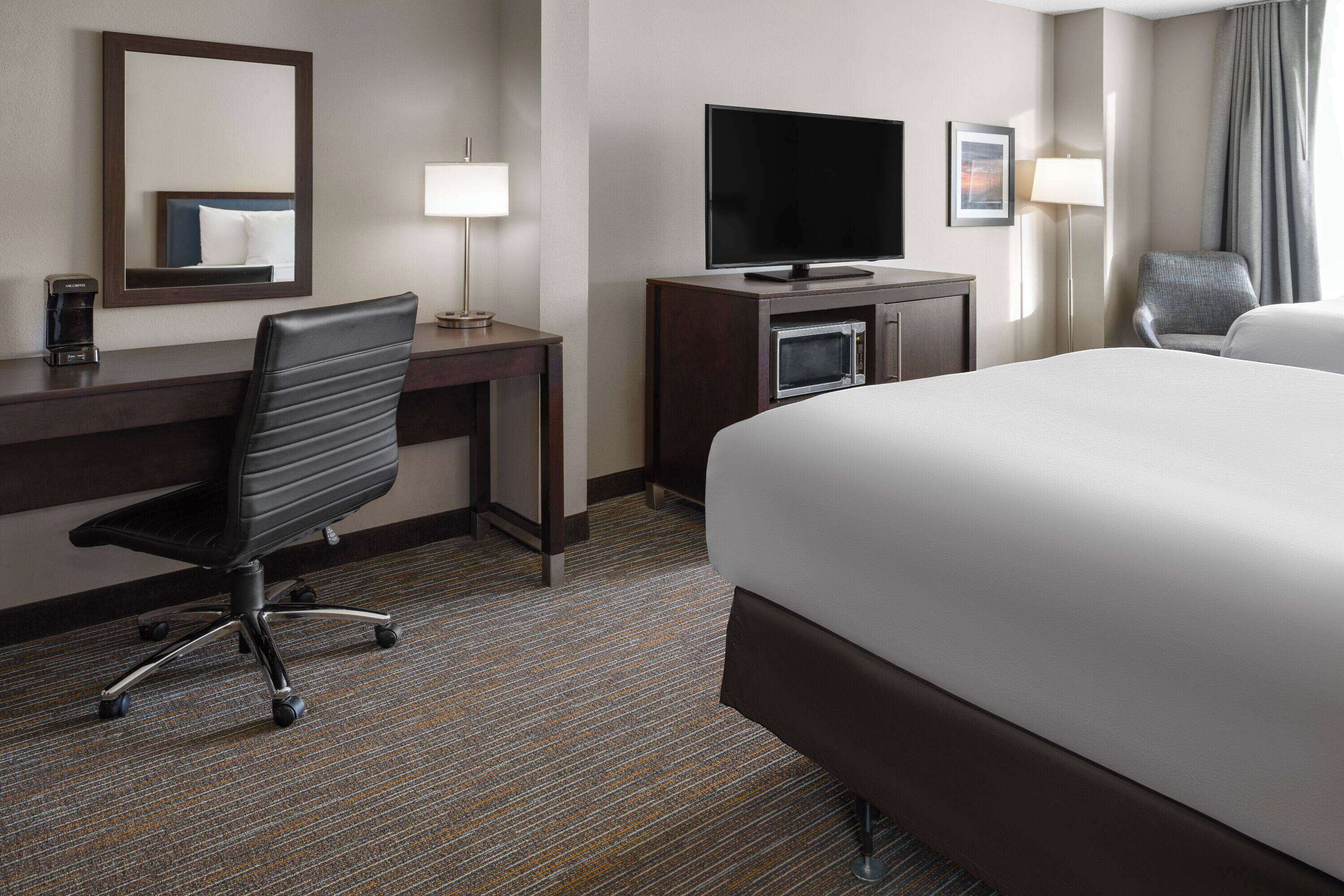 A hotel room with a large bed, a desk with a flat-screen TV, a chair, a coffee maker, and a lamp.